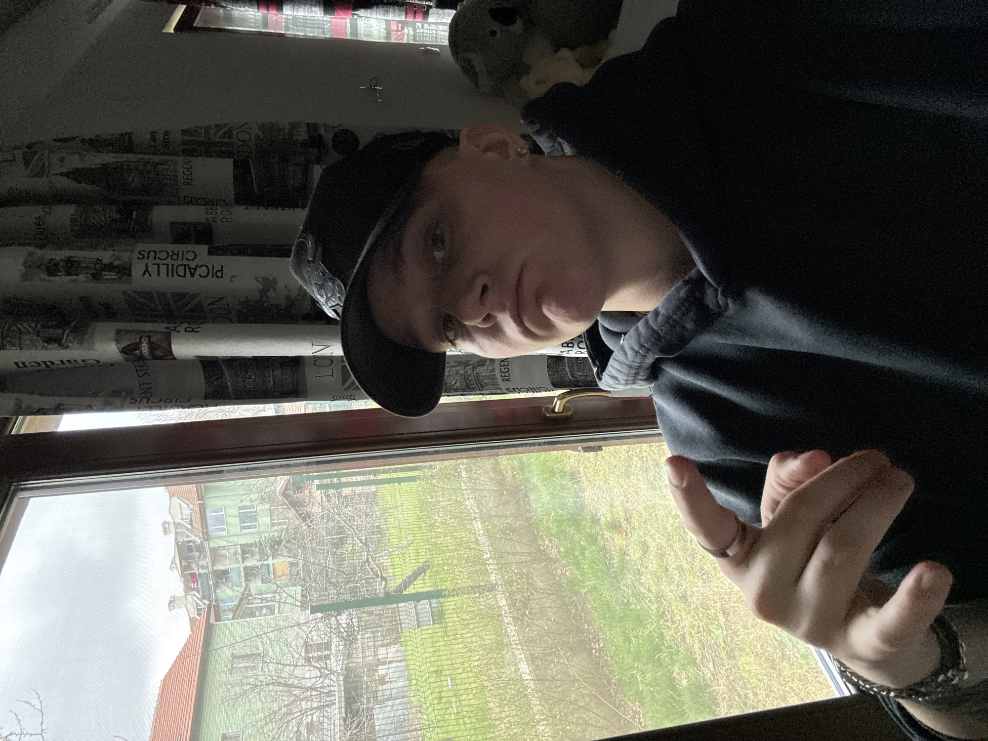 A person wearing a black hoodie and a black baseball cap is posing for a photo indoors, presumably near a window. The window overlooks a building and a grassy area, suggesting an urban setting. The interior wall near the window is covered with a newspaper-like design featuring text such as "PICADILLY CIRCUS" and "LONDON", although the wallpaper is a decorative motif only. The person is making a hand gesture that could be "I love you" in sign language. The angle of the image is tilted, possibly taken from a phone. Without more context, the location in the city of Sofia, Bulgaria, cannot be accurately determined based solely on the visible details.