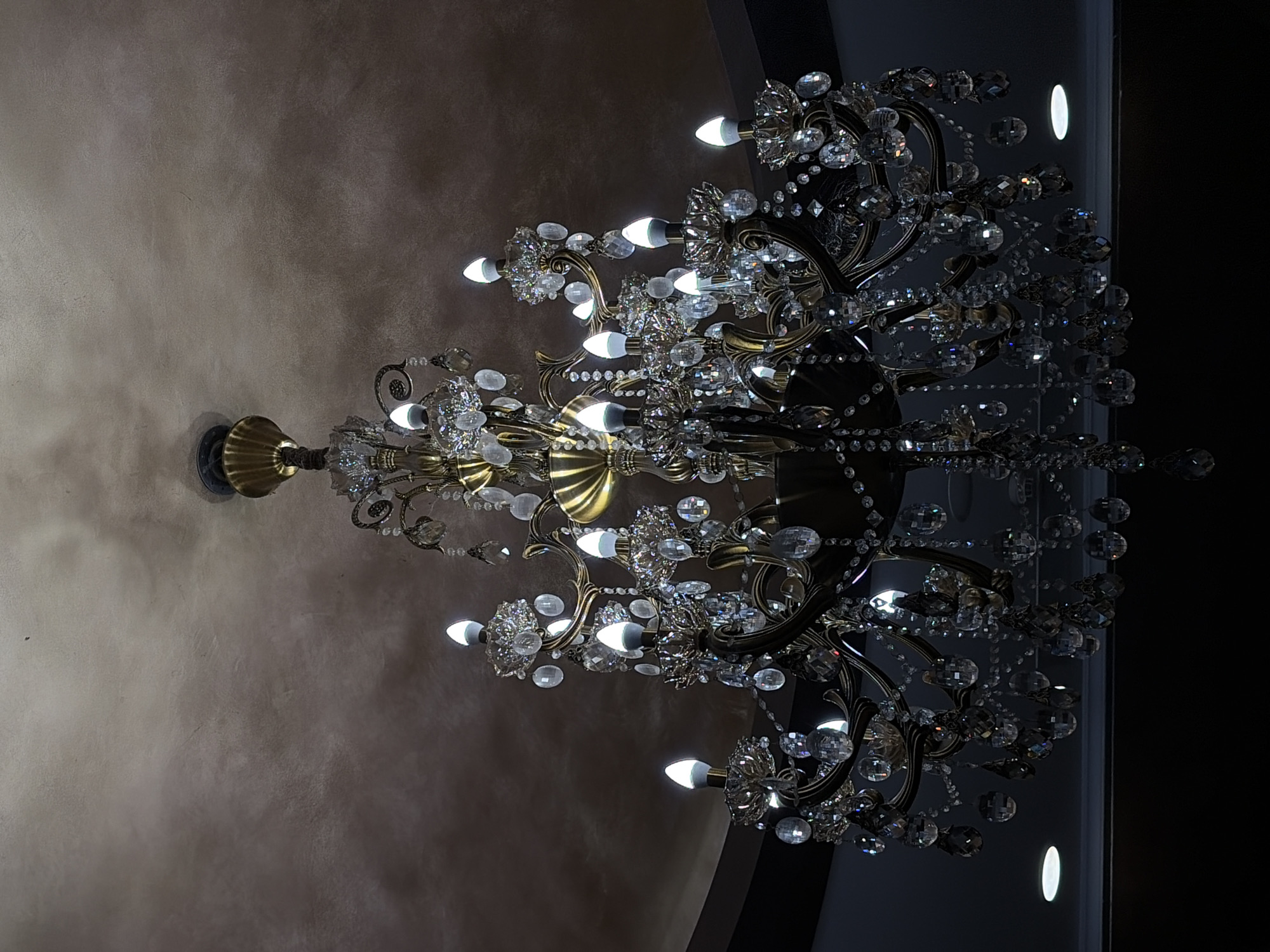A grand, ornate chandelier, crafted from brass or gold-toned metal, is the central subject of the image. It is lavishly decorated with countless sparkling clear crystal elements, including multifaceted beads, pendants, and clusters, which catch and reflect light. Numerous small, white, globe-shaped light bulbs are illuminated, contributing to the chandelier's dazzling brilliance.
The chandelier hangs from a textured ceiling, which appears to be a warm, earthy tone, possibly light brown or beige, with a stucco-like finish. To the right of the chandelier, a dark vertical surface, perhaps a wall or column, is visible, featuring a small, oval-shaped light fixture that emits a soft glow.
The scene is indoors, likely in the evening or at night, given the prominence of artificial light sources and the dim background. The elaborate design of the chandelier and the refined ceiling suggest an upscale or luxurious interior setting, consistent with venues found in locations like Budva, Montenegro. There are no people visible, and no specific activity is taking place. The image conveys an atmosphere of elegance and grandeur. No text is visible.