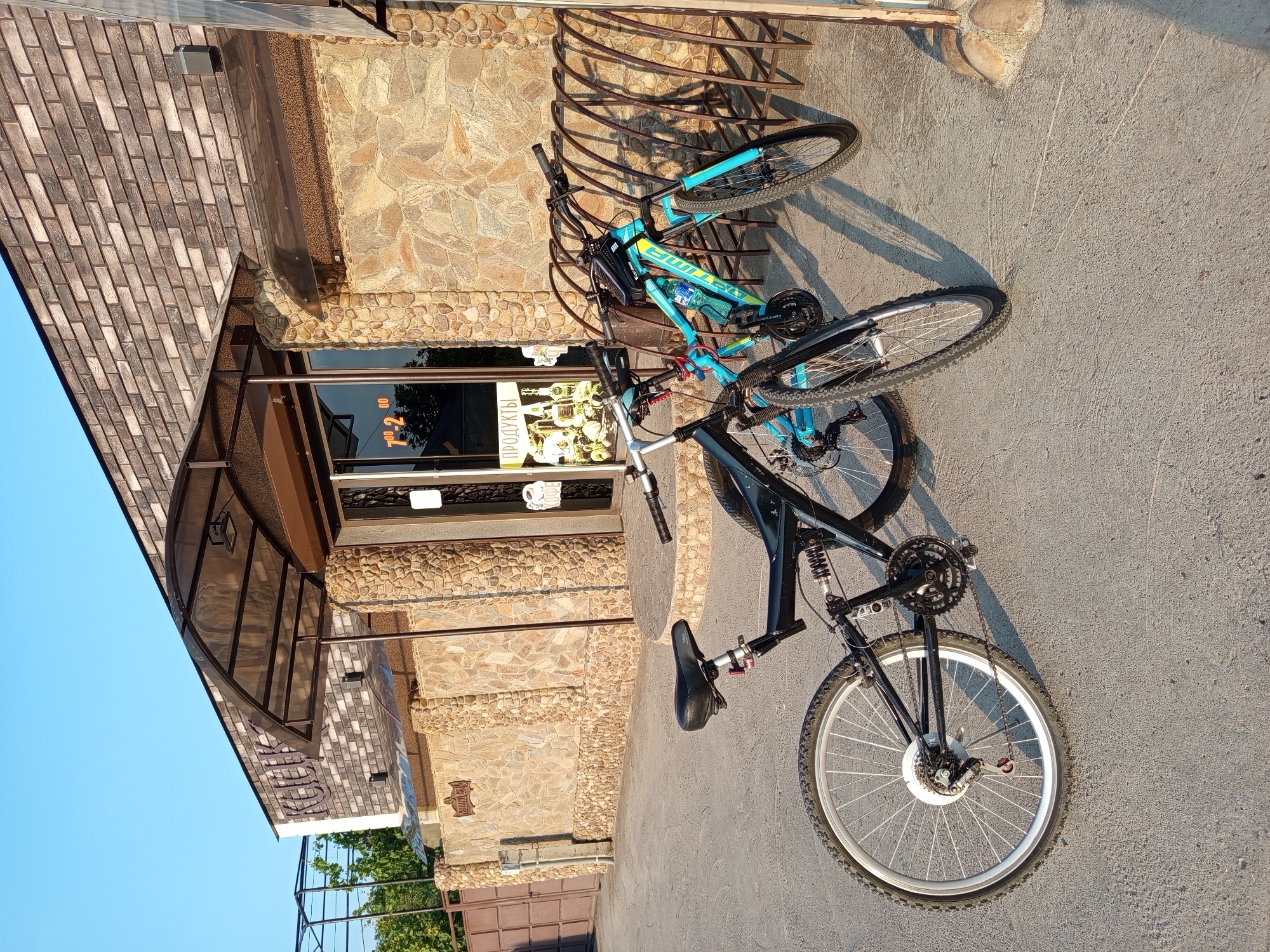 The image shows two bicycles parked in front of a building with stone and brick facade, possibly a shop or restaurant, in Kitskany, Moldova. The building has large glass doors and windows. Awnings extend over the entrance. A bicycle rack is visible to the right of the bicycles. The ground is paved with concrete. Trees and a fence are visible in the background. The sky is clear and blue. One bicycle is black and has a full suspension system, while the other is blue and yellow. The lighting suggests it is daytime, possibly late afternoon.