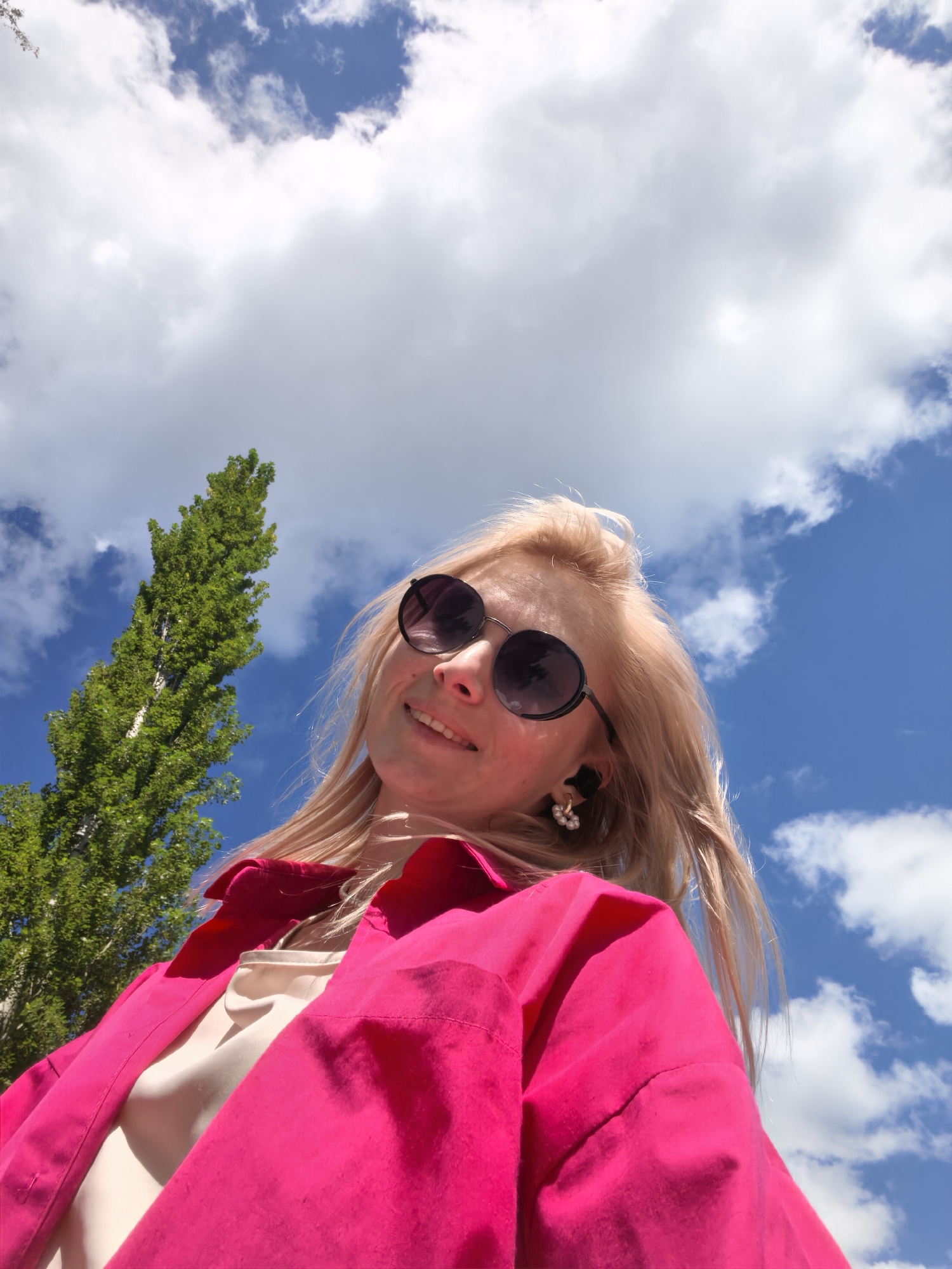 A low-angle shot captures a young woman with blonde hair, wearing round sunglasses and a bright pink shirt, against a backdrop of a vibrant blue sky filled with white, fluffy clouds and a tall, green tree on the left. The woman is smiling and looking upwards. The image appears to be taken outdoors in Sterlitamak, Russia.