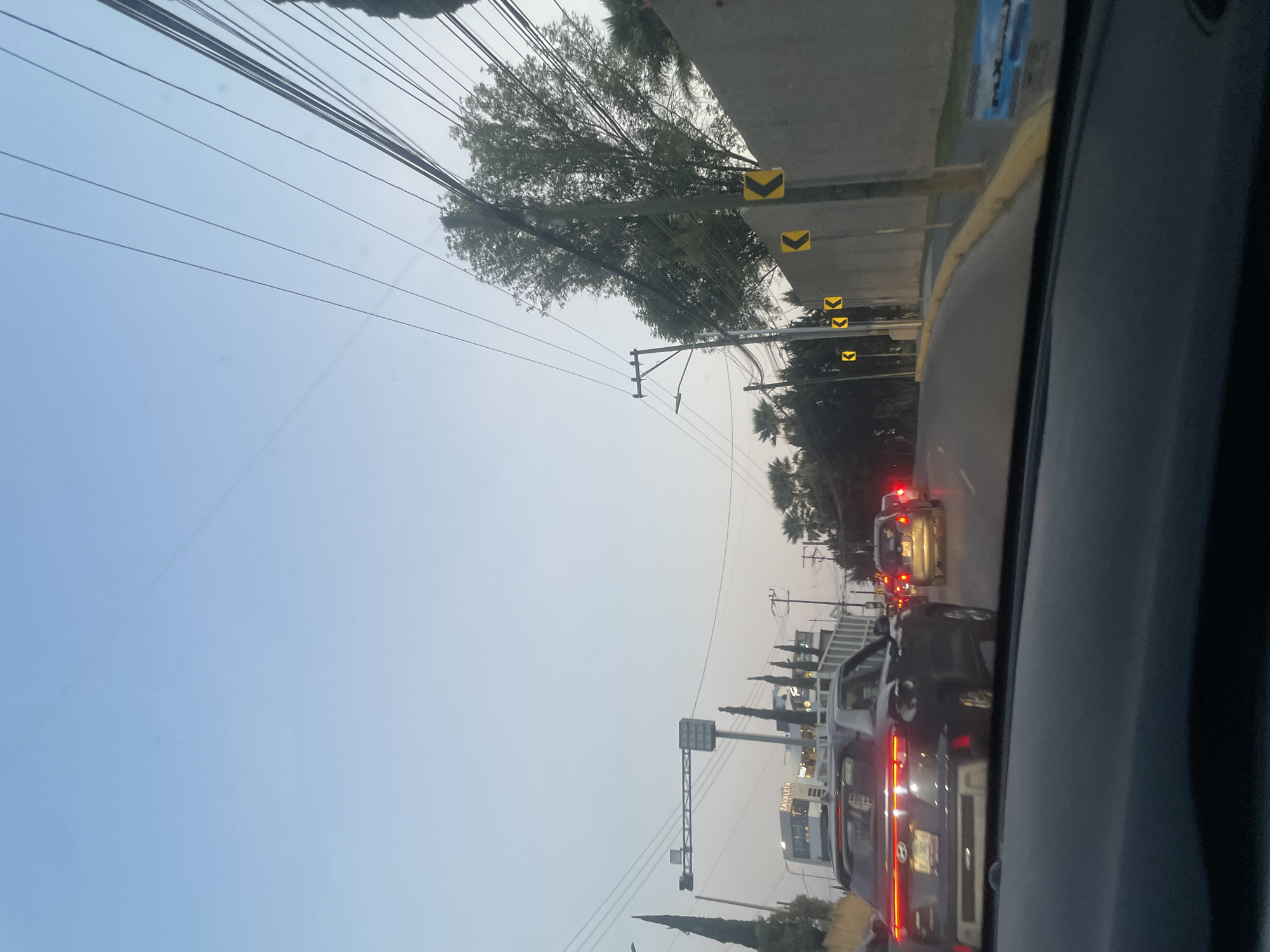 The image shows a traffic jam on a road in Puebla, Mexico. In the foreground, a car's interior is visible, with the dashboard and part of the windshield in view. The road is congested with cars, with visible taillights and brake lights. A commercial area with buildings is to the right, with a sign that reads "Zavaleta." Palm trees and other vegetation are visible. Overhead, there are power lines and a bridge with road signs. The sky is overcast, suggesting early morning or late afternoon light.