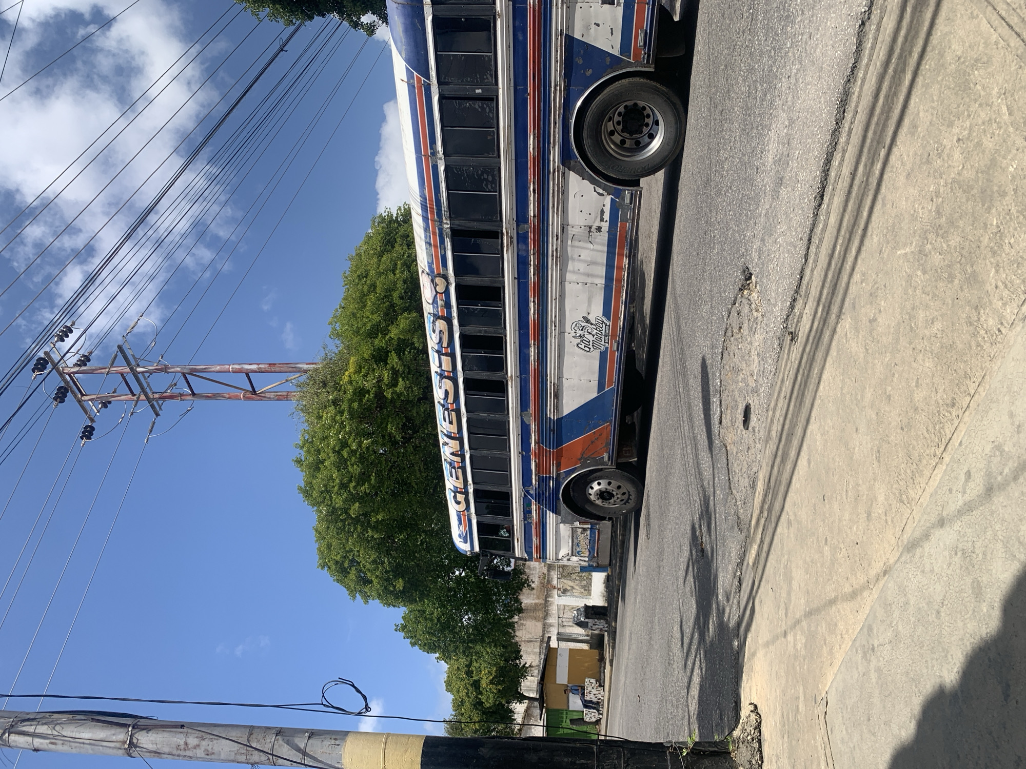 The image shows a colorful, weathered bus with the word "GENESIS" visible on its side, parked on a street in Santa Ana, Venezuela. The bus has a blue, white, and red striped design with orange accents. There is a small graphic of a monkey wearing sunglasses and the text "Gao Monkey" on the lower side of the bus. The bus is positioned on the right side of the frame, on an asphalt road. To the left of the bus is a sidewalk and a utility pole with visible wires. Behind the bus, there are trees and a low building with a yellow and green facade, where a person in a blue shirt is seated. The sky is clear blue with some white clouds, and power lines crisscross the upper portion of the image. The road surface appears aged and shows some signs of wear, including potholes.