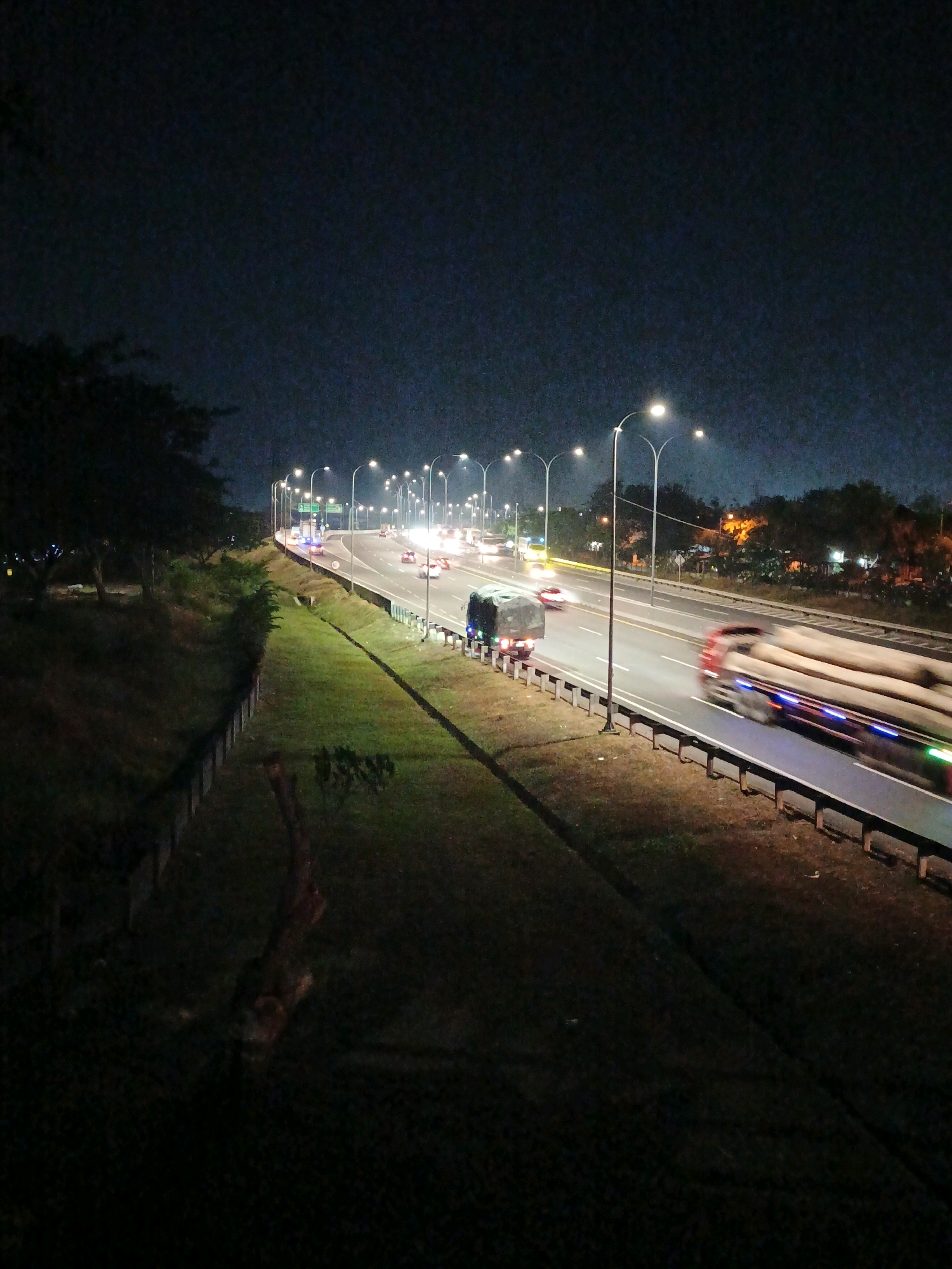 The image depicts a night scene on a multi-lane highway in Kludan, Indonesia. The highway is illuminated by a series of streetlights, creating a bright pathway through the darkness. Several vehicles are visible on the road, including cars and trucks. One truck in the foreground is carrying a large load of logs and is in motion, causing a blur due to the long exposure. Another truck, covered with a tarp, is visible in the left lane. The highway is bordered by grassy embankments and trees. The sky is dark, indicating nighttime.
