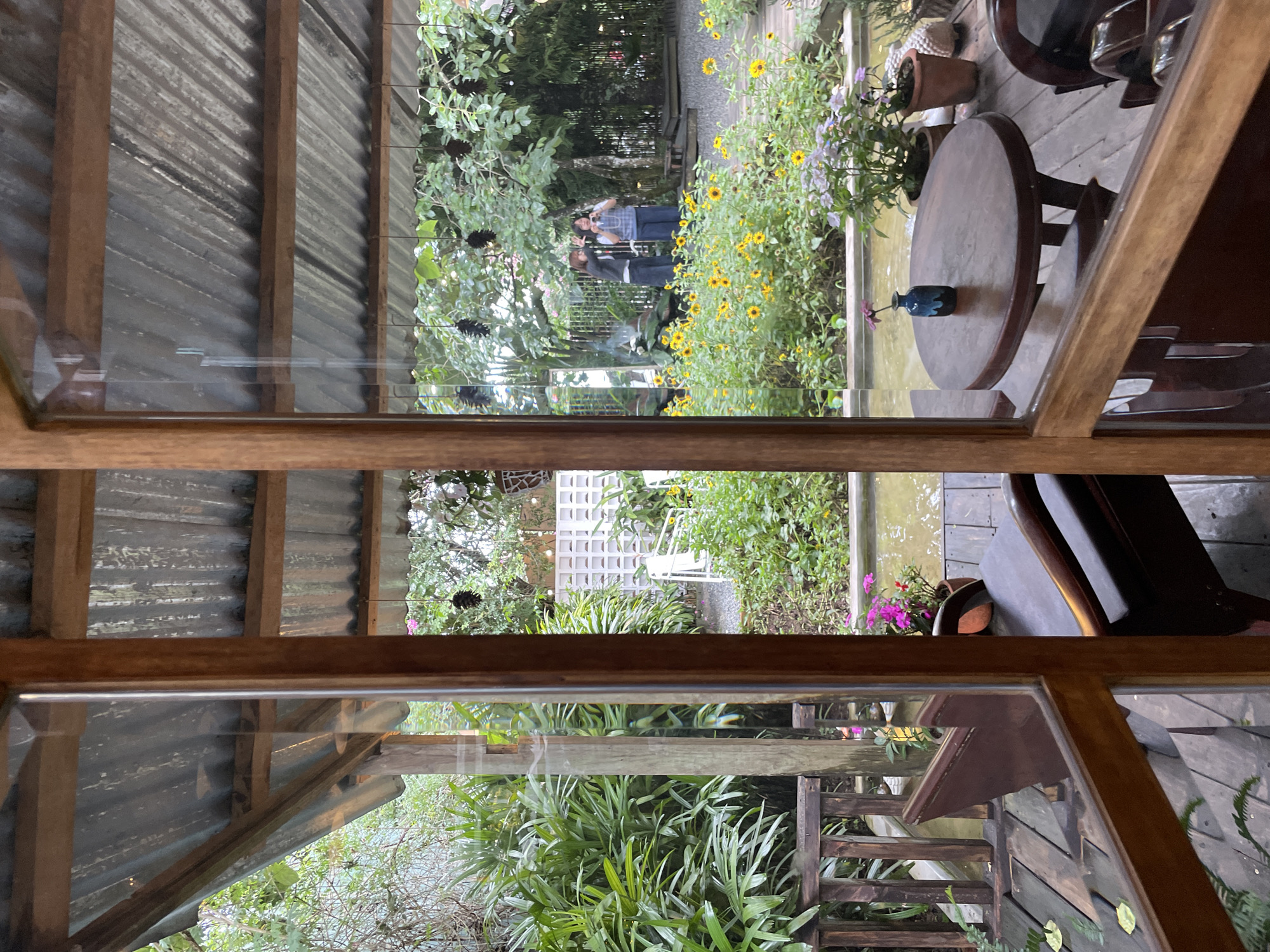This is an exterior view through a wooden-framed window or door of a covered outdoor seating area, likely a cafe or restaurant in Da Lat, Vietnam. The scene is dominated by lush greenery and a small garden. In the foreground, on a dark wooden deck, are two dark wooden armchairs and a round wooden table with a small blue vase holding a single purple flower. Beyond the seating area, a shallow body of water or pond is visible, with more plants lining its edges. Further back, a garden bursts with yellow sunflowers and other flowering plants, interspersed with trees. Two women are visible in the background, standing among the flowers; one is holding up a phone, possibly taking a picture. Behind the garden is a decorative white lattice wall and more trees. The roof of the covered area is made of corrugated metal supported by wooden beams. The overall atmosphere is natural, tranquil, and slightly rustic.