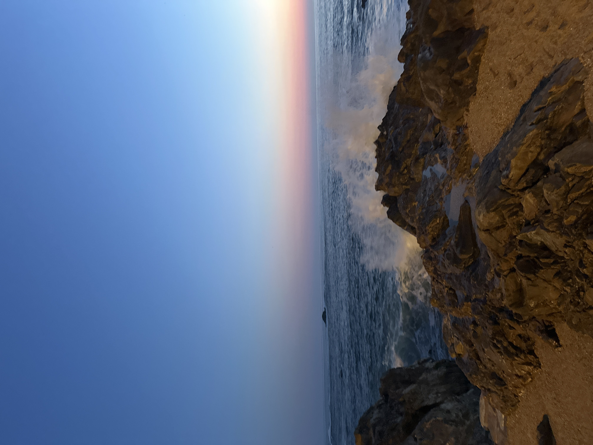 The image displays a coastal scene at twilight along the Atlantic coast in Foz do Douro, Porto, Portugal. The sky transitions from a deep blue overhead to a lighter blue, then to soft pink and orange hues at the western horizon, indicating sunset. The dark blue-grey ocean exhibits moderate wave activity. A large white wave is captured mid-crash against a formation of dark, rugged rocks in the mid-right foreground. A smaller, dark, low-lying rock is discernible on the distant horizon towards the left-center. The immediate foreground consists of extensive dark, irregular rock outcrops, appearing wet from sea spray, with visible small pools of water. A patch of light brown gravel or sand occupies the extreme lower right portion of the frame. No human figures, animals, or distinct man-made structures are present within the captured scene.