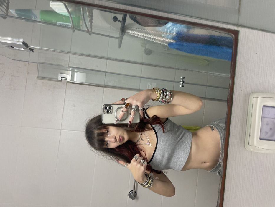 A young woman poses in front of a mirror in a bathroom. She wears a gray crop top and sweatpants, adorned with multiple rings and bracelets. Her long brown hair has red highlights and bangs that cover her forehead. She holds a smartphone in her right hand, taking a selfie. The reflection shows a tiled bathroom, a shelf with toiletries, and a thermostat on the wall.