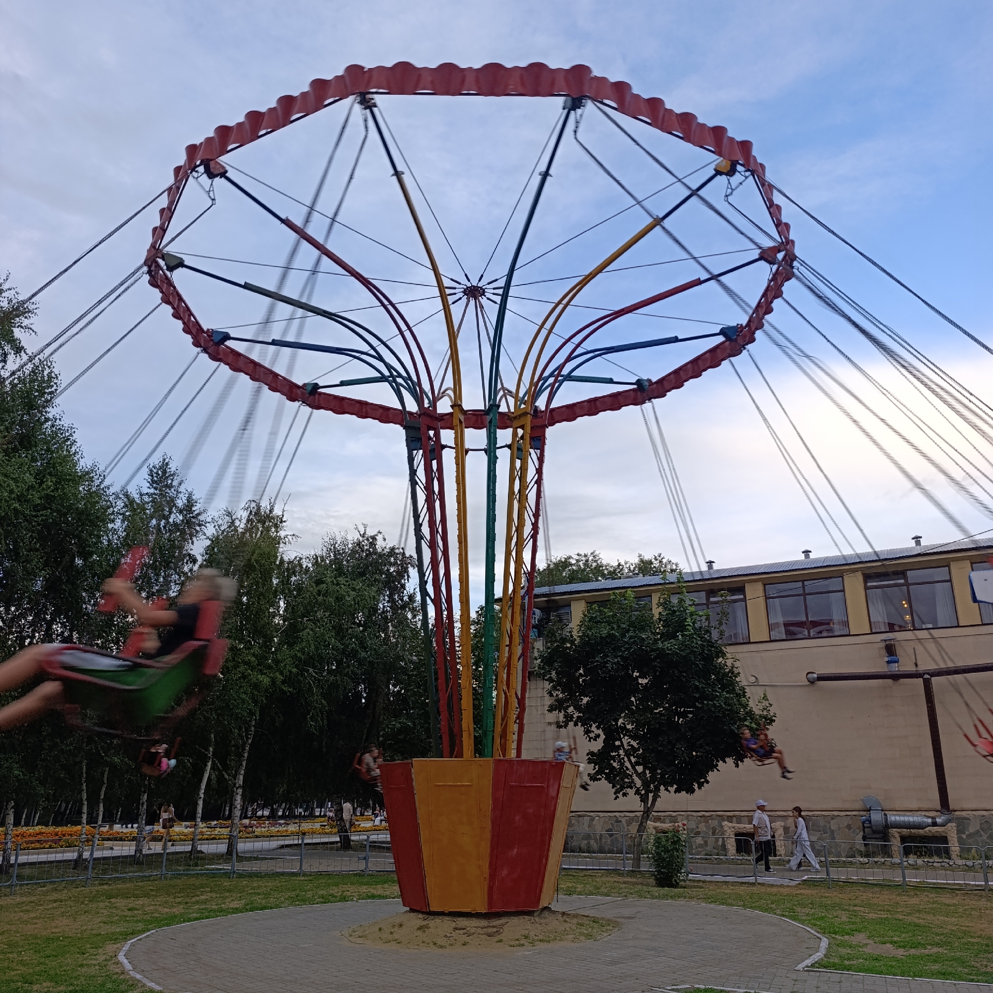 The image depicts a carousel swing ride in an amusement park located in Kostanay, Kazakhstan. The ride features a central column with colorful poles (red, yellow, and green) supporting a circular canopy. Several swings are attached to the canopy by chains, and some are in motion, with blurred figures of people visible on them. The ride is situated on a paved circular area surrounded by grass and trees. In the background, a building with large windows and a yellow facade is visible, along with more trees and a fence. Two individuals are walking in front of the building, near the fence. The sky is partly cloudy, with a mix of blue sky and white clouds. The overall scene suggests a recreational area within the city.