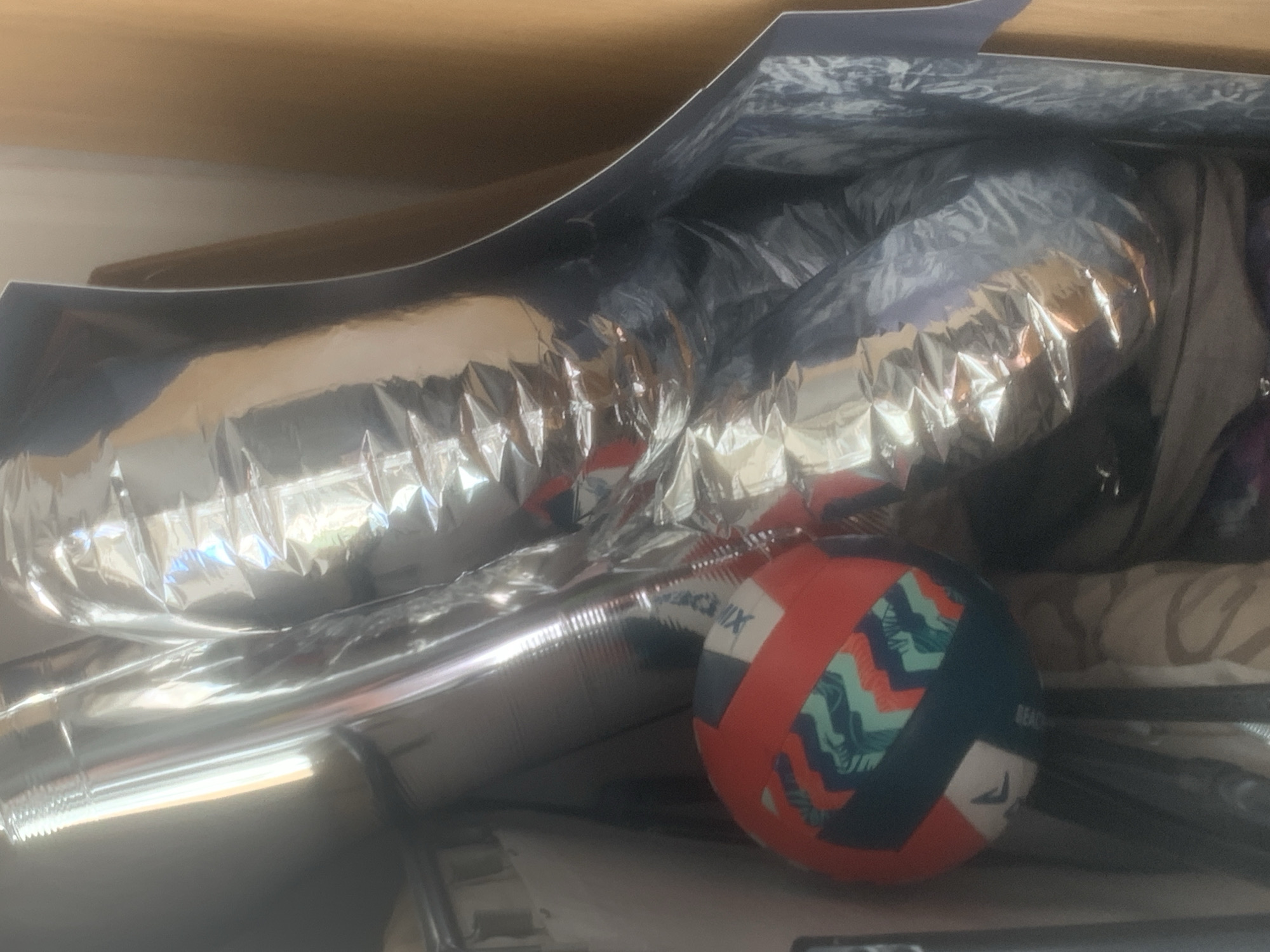 The image shows a cluttered storage space, possibly indoors. The dominant feature is a large, crumpled piece of shiny, reflective material resembling aluminum foil or mylar packaging. Nestled among the crumpled material is a small, multi-colored ball, predominantly orange, blue, and white, with geometric patterns. The background is filled with indistinct objects, suggesting a crowded environment. There is no evidence to connect the image to the city of Novoe Devyatkino, Russia.