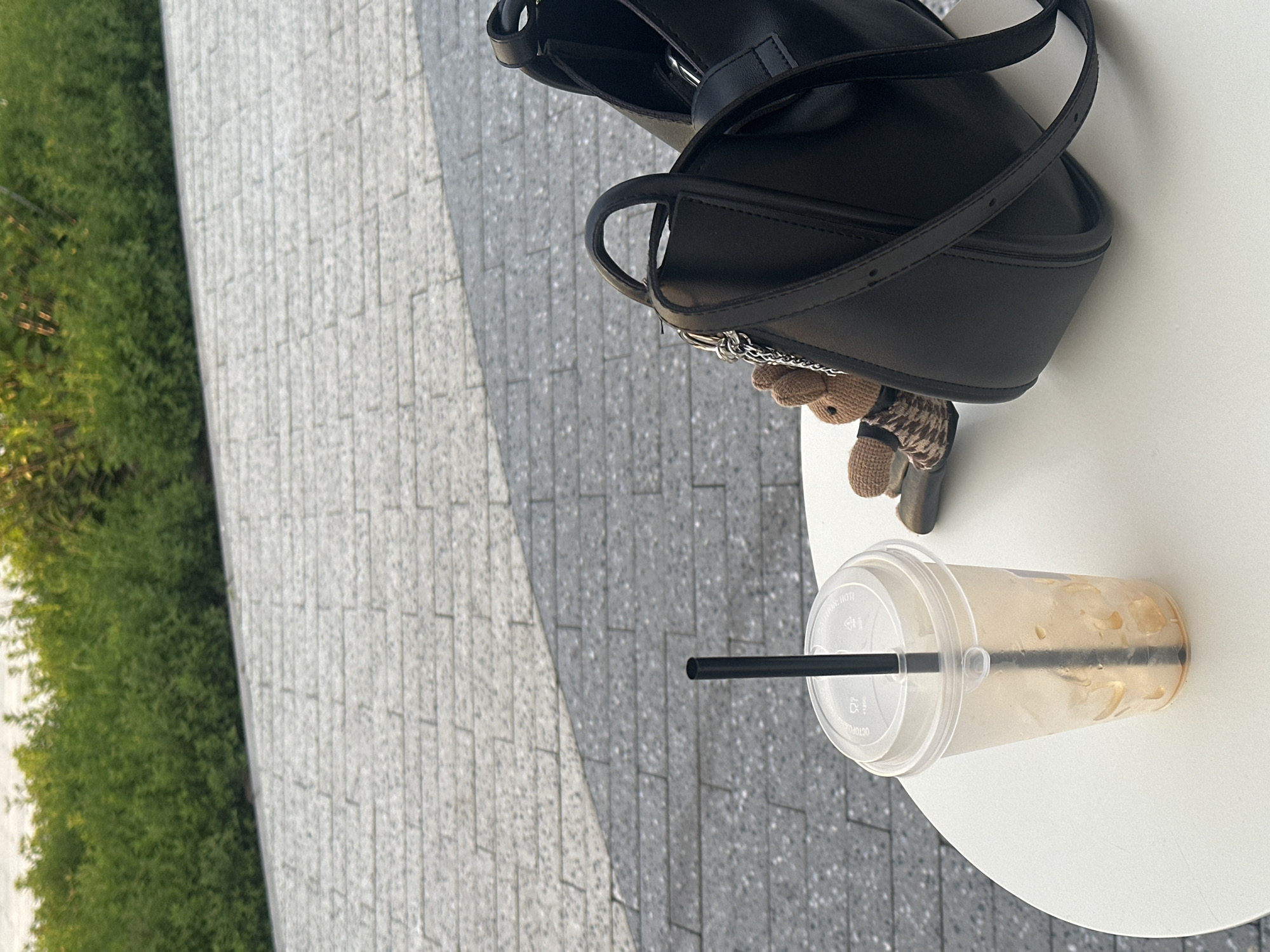 The image depicts a partially consumed iced coffee in a plastic cup with a black straw on a round white table. Adjacent to the drink is a black leather purse with a brown and black patterned keychain. The table sits on a gray brick-paved surface, and lush green foliage is visible in the background. The setting appears to be outdoors. The location cannot be confirmed based on the image.