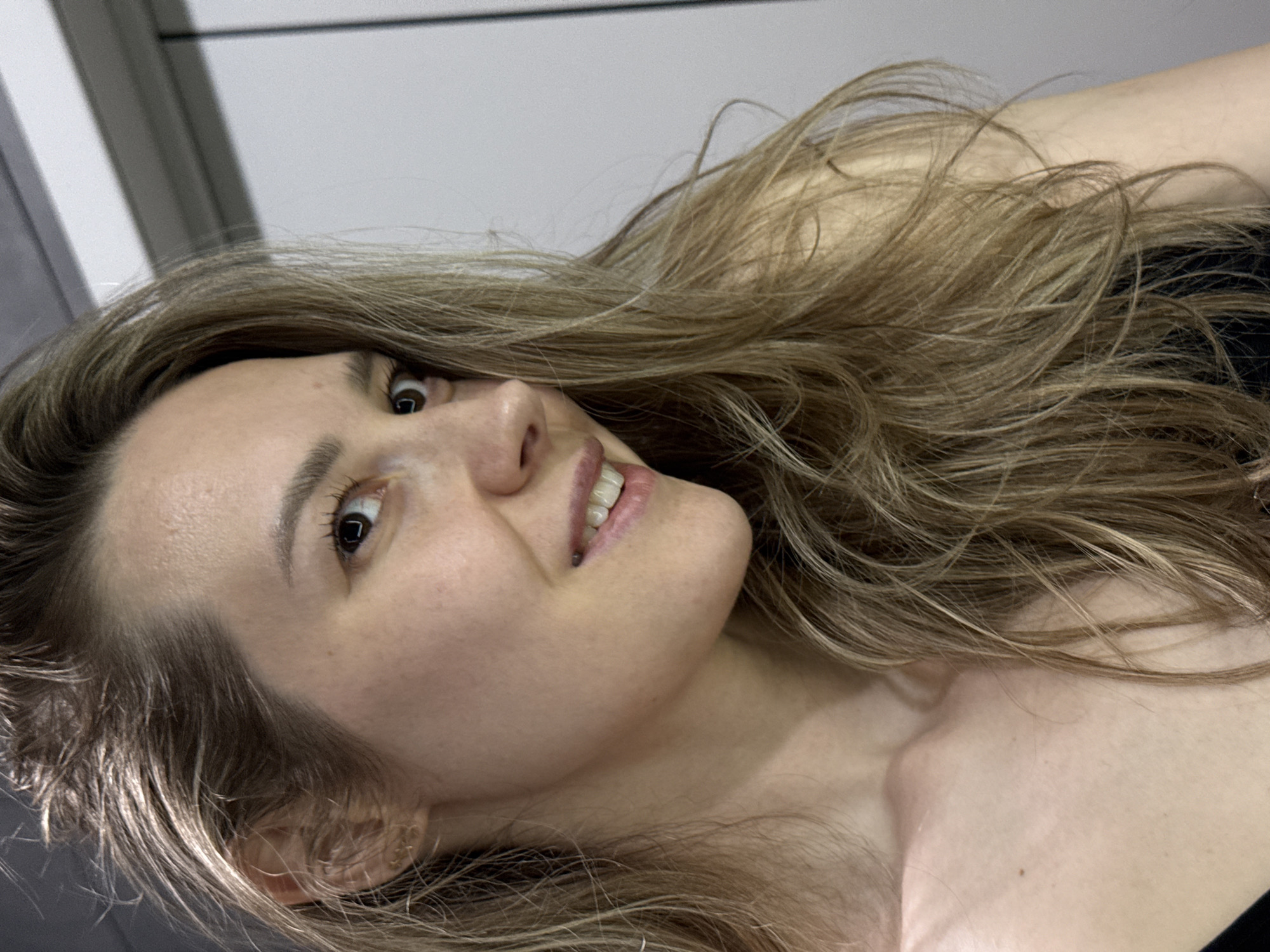 A light-skinned woman with light brown hair, partially in focus, is the primary subject. The woman is facing the camera with a slight smile. Her hair is long and wavy. The background features a plain, light gray surface, possibly a wall, with a darker gray element on the left side. The setting cannot be reliably determined to be in the city of Chaikovsky, Russia.