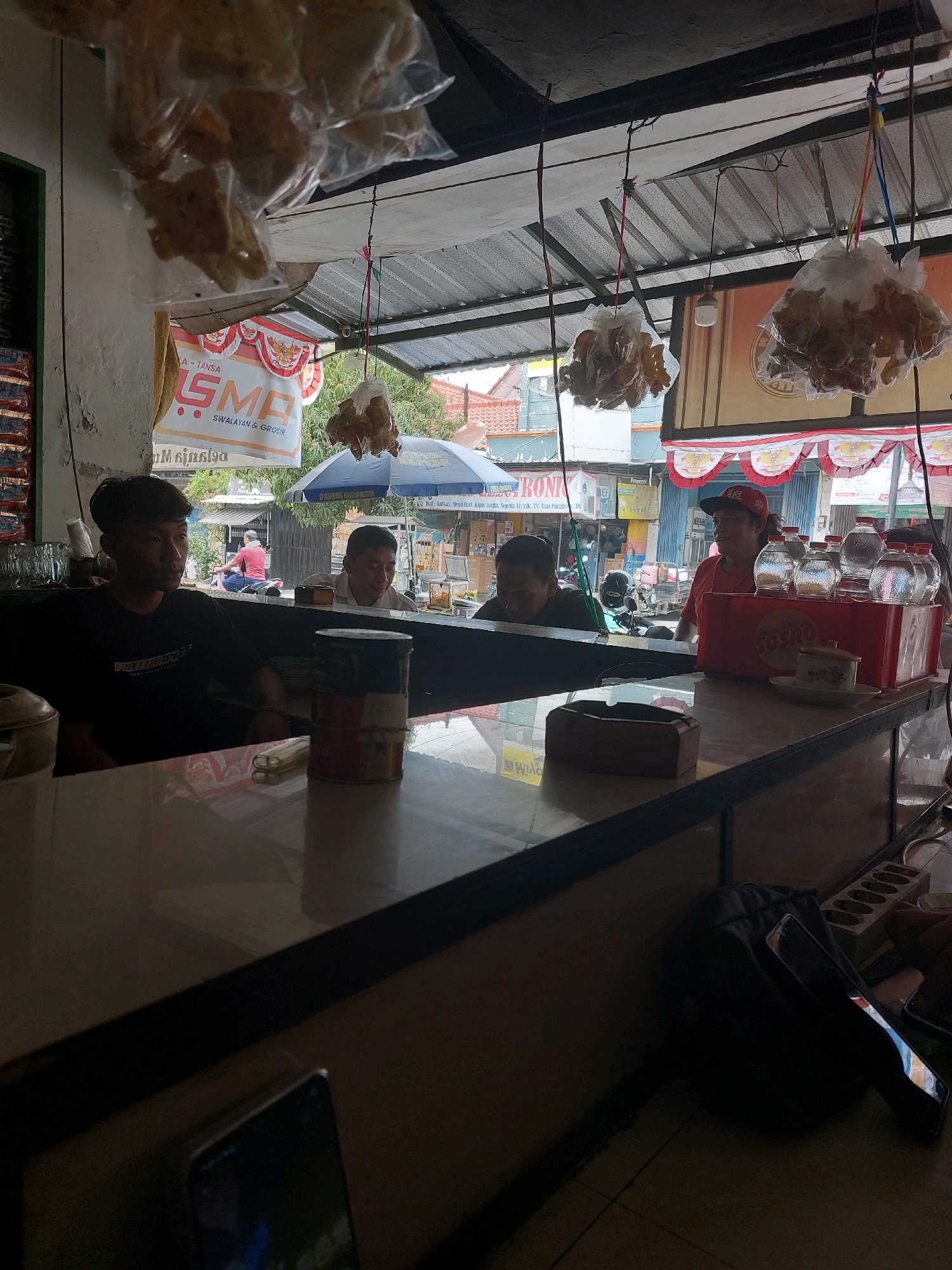 The image depicts an outdoor roadside stall in Sumuragung, Indonesia, possibly a warung. The scene is dimly lit with natural light filtering through a corrugated metal roof structure. In the foreground, a long counter is visible, made of a light-colored material with a dark edge. On the counter are several items including a wooden ashtray, a large cylindrical container with red, blue, and yellow bands, and what appears to be a small ceramic dish. Behind the counter, three individuals are partially visible. A young man in a black t-shirt is seated on the left, looking to his left. In the middle, a man in a white collared shirt is looking down. On the right, a person wearing a red t-shirt and a red baseball cap is standing and looking to their left. Behind the counter and the people, a row of glass bottles is arranged on a red tray. In the background, the street is visible with buildings, trees, and a person on a motorcycle. Several plastic-wrapped bags containing food items are hanging from the roof structure. A blue umbrella is also visible in the background, as well as various signs and products typically found in Indonesian roadside stalls. A dark backpack and a mobile phone are on the floor in the lower right portion of the image.