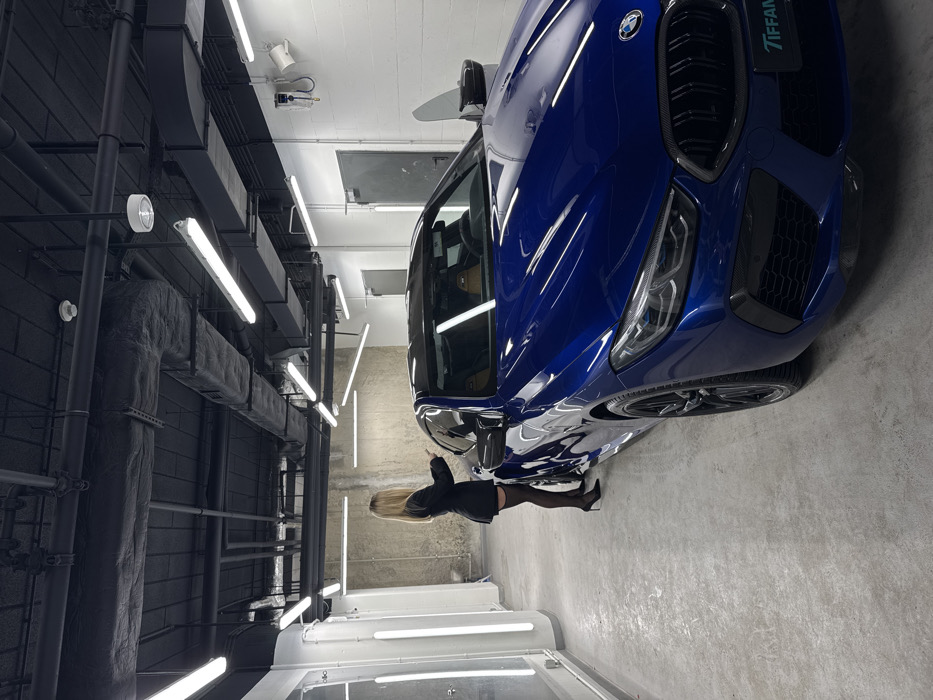 The image depicts a professional car detailing or service bay in Warsaw, Poland. A dark blue BMW M8 coupe, featuring a light brown leather interior, black wheels, and a visible "TIFFANY" plate on its front bumper, is centrally positioned, angled slightly towards the left. The driver's side door of the vehicle is open.
A single female individual is standing on the driver's side of the BMW, viewed from behind. She has long blonde hair and is wearing a black blazer, black sheer stockings, and black high heels. Her right arm is raised and bent, appearing to interact with the car's interior or the open door. Her left hand is also slightly raised.
The environment features a smooth, light gray concrete floor. The walls consist of white painted cinder blocks on the right, exposed concrete at the back, and white panels with vertical linear lights on the left. The ceiling is painted black, exposing industrial elements such as HVAC ducts, pipes, and electrical conduits. Numerous horizontal linear LED light fixtures are suspended from the ceiling, providing bright and uniform illumination. A wall-mounted utility unit with hoses is visible on the right wall, and two metal doors are present on the back wall.