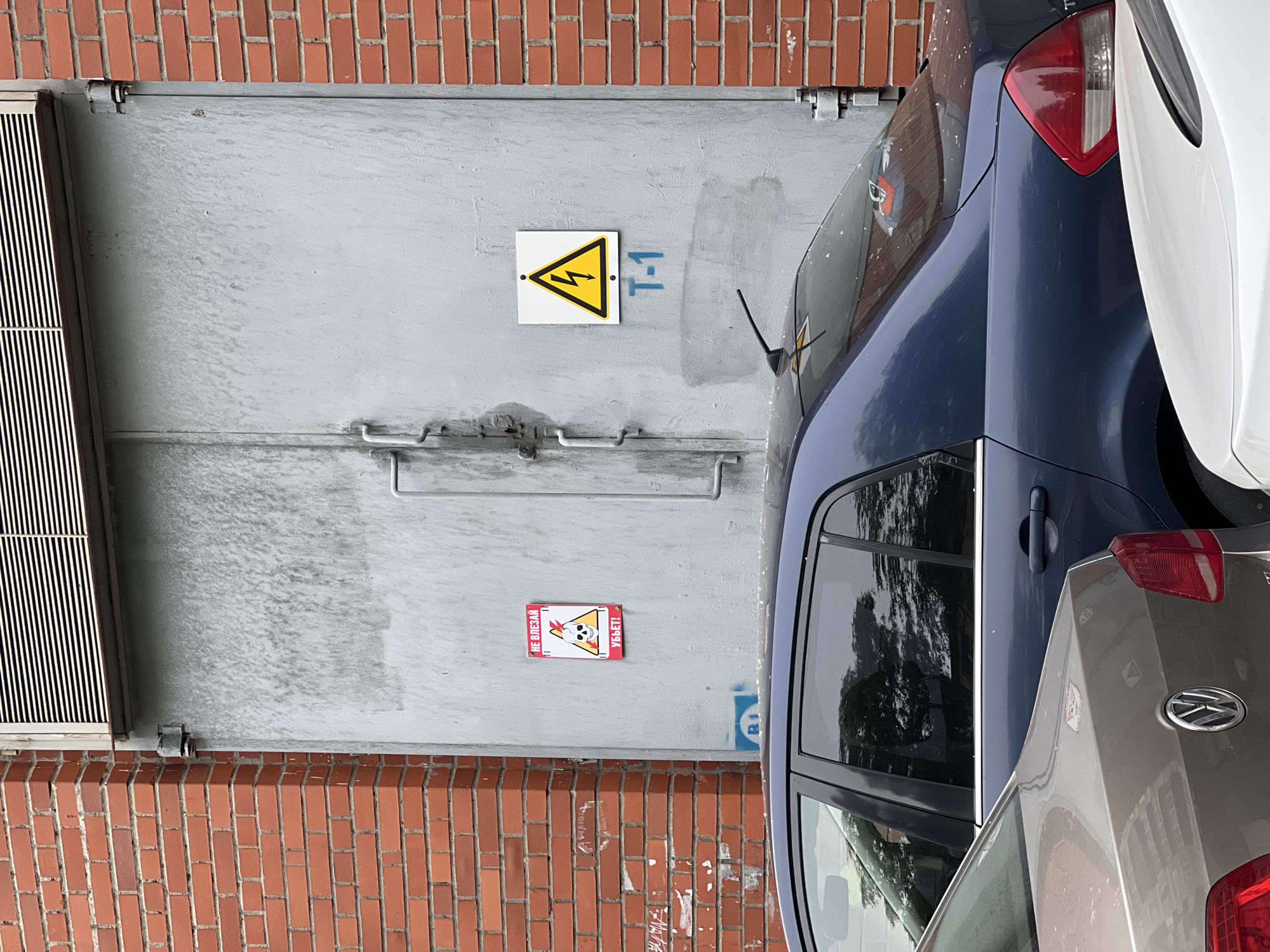 The image shows a section of a building exterior with a large, grey metal door. To the left of the door, a red brick wall is visible. On the metal door, there are two warning signs. The sign on the left is a red and white rectangular sign with a skull and lightning bolt symbol, and text in Russian that translates to "DO NOT ENTER, ELECTRICITY WILL KILL YOU!". The sign on the right is a white square with a yellow lightning bolt symbol inside a black triangle, indicating high voltage. Below this sign, "T-1" is written in blue. In front of the door, two cars are parked, partially obscuring the lower part of the door. The first car is a dark blue sedan, and the second car is a lighter-colored sedan in front of it. The overall environment appears to be an urban setting, possibly an industrial or utility area, given the warning signs and the nature of the building. The location is identified as Unknown, Russia.