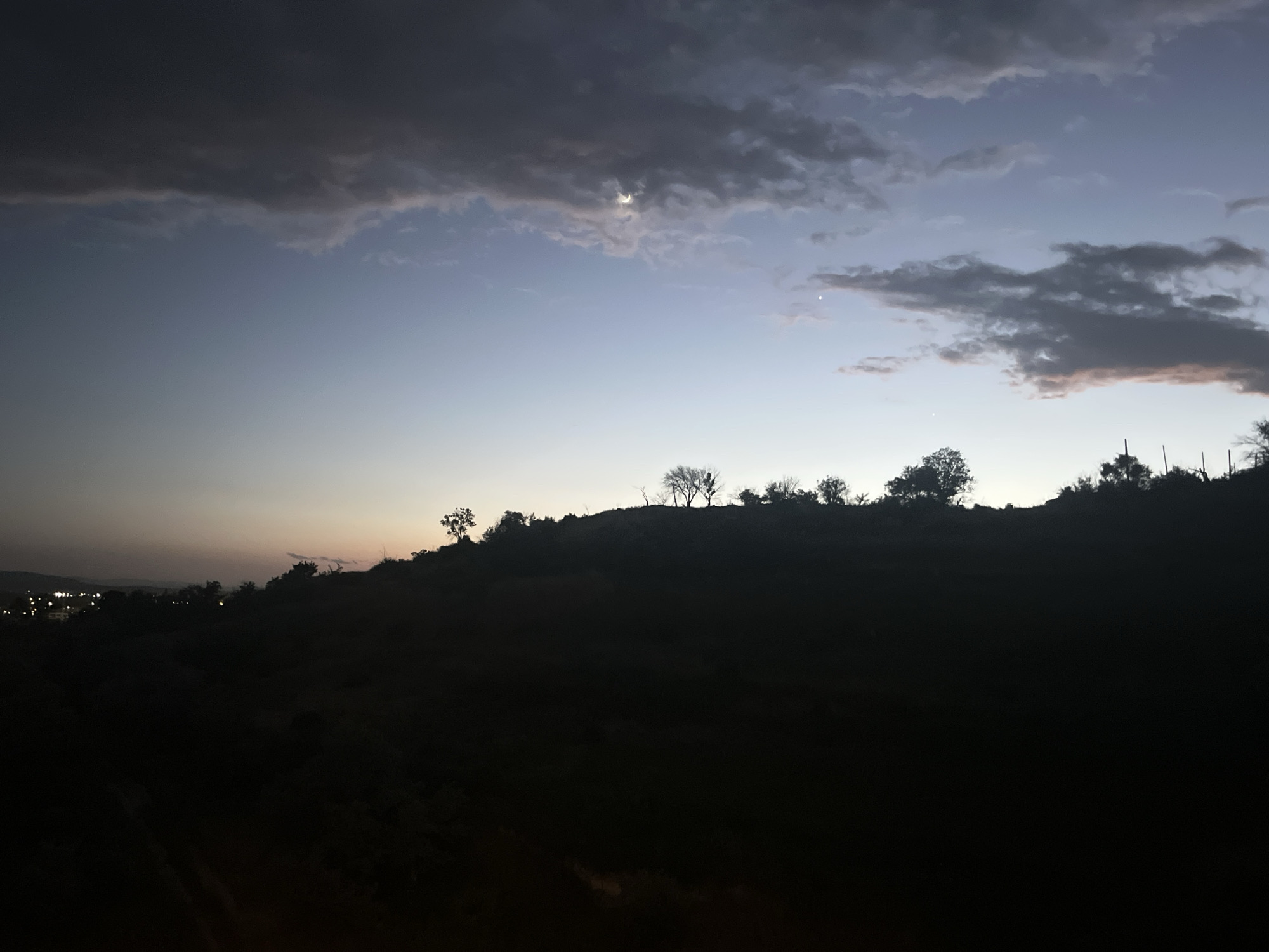 The image depicts a twilight sky over a silhouetted landscape. Dark, heavy clouds occupy the upper portion of the frame, with a sliver of the crescent moon visible through a break in the clouds. Below the clouds, the sky transitions to a pale blue, fading to a lighter hue near the horizon. A dark, undulating hillside dominates the lower half of the image, with the silhouettes of scattered trees and vegetation visible along its crest. In the distance, on the left side of the frame, faint lights suggest a populated area. The overall impression is one of a serene, natural landscape at dusk, with the encroaching darkness creating a dramatic contrast against the pale sky. The location of Varna, Bulgaria, is not discernible from the visual information provided in the image.