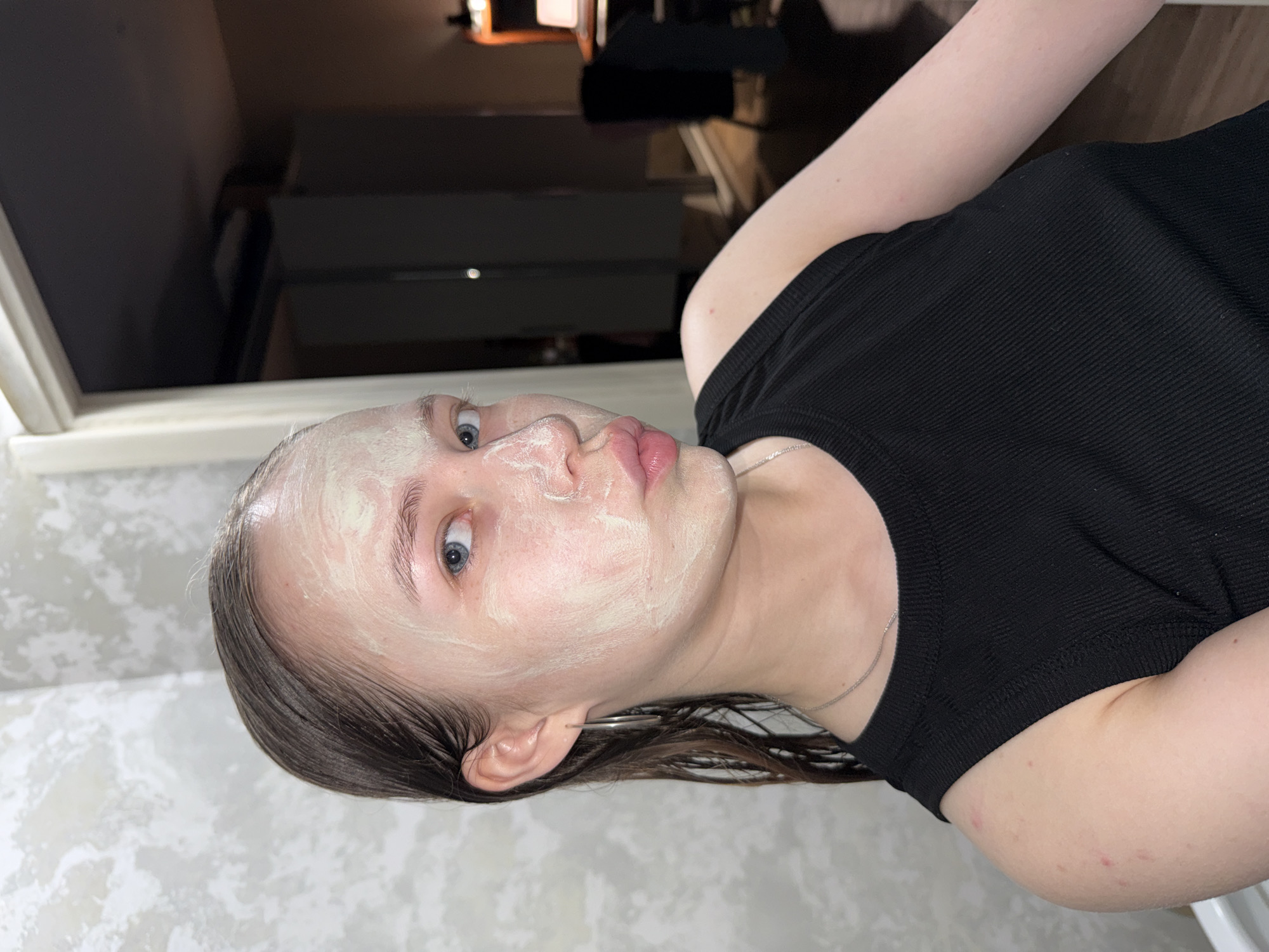 A young Caucasian female with wet brown hair and blue eyes, wearing a black ribbed tank top and silver hoop earrings, is looking at the camera with her lips pursed. She has a face mask applied to her forehead, cheeks, and nose. She is indoors, with a textured grey wall behind her to the left and a doorway leading to a dimly lit room with furniture visible on the right. There are no identifiable landmarks suggesting the location is in Chaikovsky, Russia.