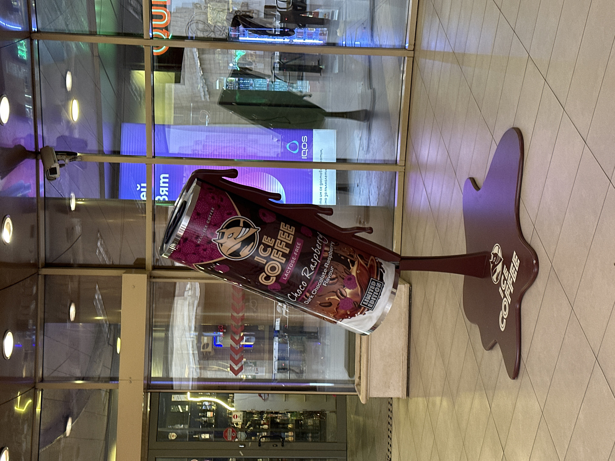 This media file captures an indoor promotional display for "HELL ICE COFFEE" in Sofia, Bulgaria. The main subject is a large, maroon-colored replica of an ice coffee can, angled as if pouring. The can's label is prominent, featuring a devil-like logo and text that reads "HELL ICE COFFEE," "ICE COFFEE," "LACTOSE-FREE," "Choco Raspberry," "Dark Chocolate-Raspberry Flavour," and "LIMITED EDITION." The "poured" liquid forms a dark, irregular shape on a matching maroon stand, which also bears the "ICE COFFEE" logo.
The display rests on a light-colored tiled floor with a diagonal pattern. Behind it, a large glass window or partition reflects and reveals elements of the indoor environment. Through and reflected in the glass, several illuminated signs are visible. One sign shows Cyrillic text, including fragments like "Зят" and a phrase indicating age restriction: "та им се доставя амо за пълнолетн" (delivered to them only for adults). Another reflection clearly shows the "IQOS" logo and the partial word "котин," likely referring to nicotine. A white security camera is mounted behind the glass, also reflected. Various light sources, including circular recessed lights and colorful artificial lighting, illuminate the scene, suggesting an evening or perpetually lit indoor setting within a commercial building, possibly a shopping mall. The image is a static shot of this elaborate product advertisement.