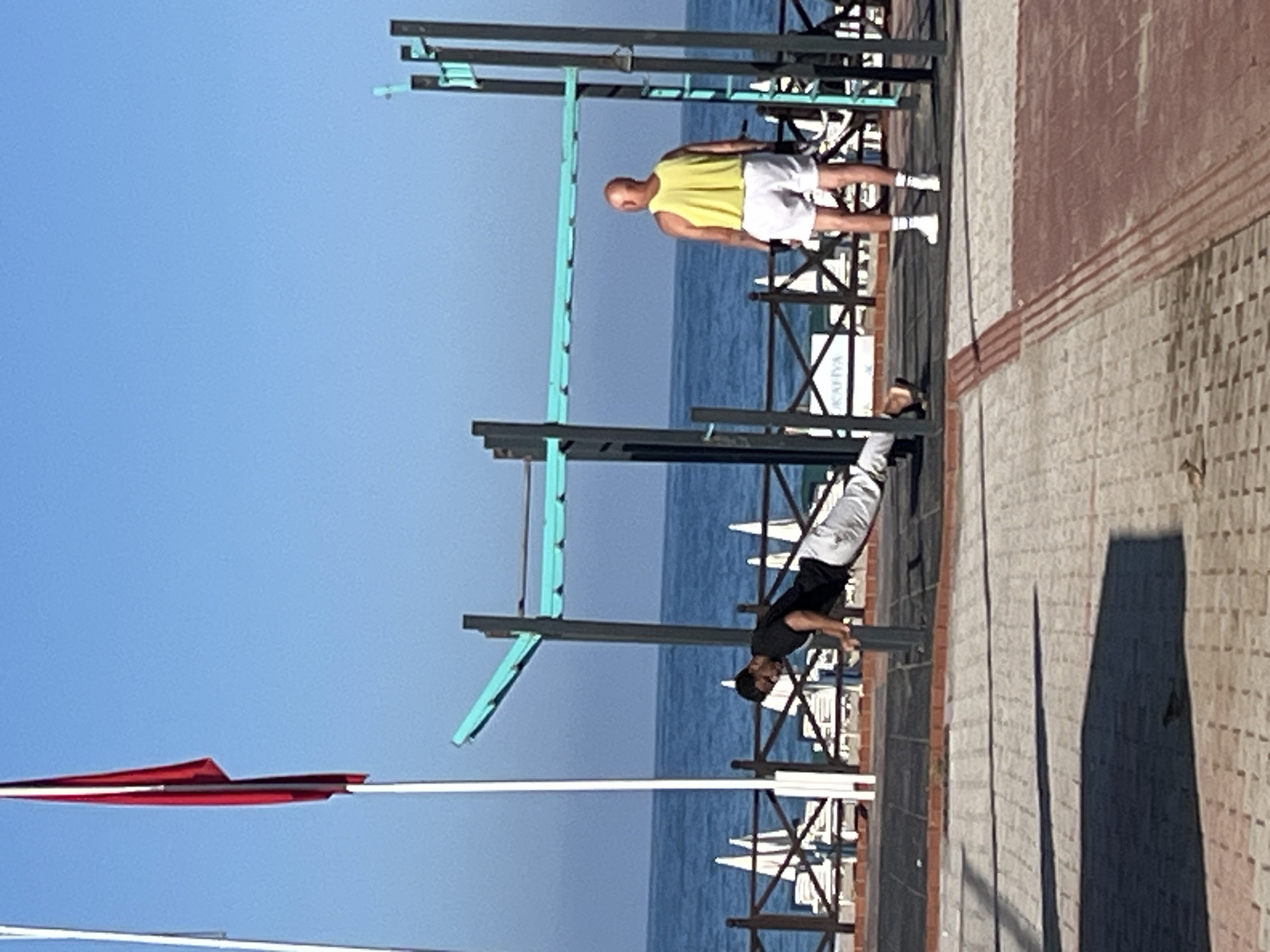 The image depicts an outdoor workout area in Alanya, Turkey, with two men exercising on pull-up bars. One man, wearing a yellow tank top and white shorts, is standing on the ground near the bars. The other man, dressed in a black shirt and white pants, is using the bars for a workout. The workout area is located on a paved surface next to a railing, with beach umbrellas and the sea visible in the background. A red and white flag is attached to a white pole in the foreground. The sky is clear and blue.