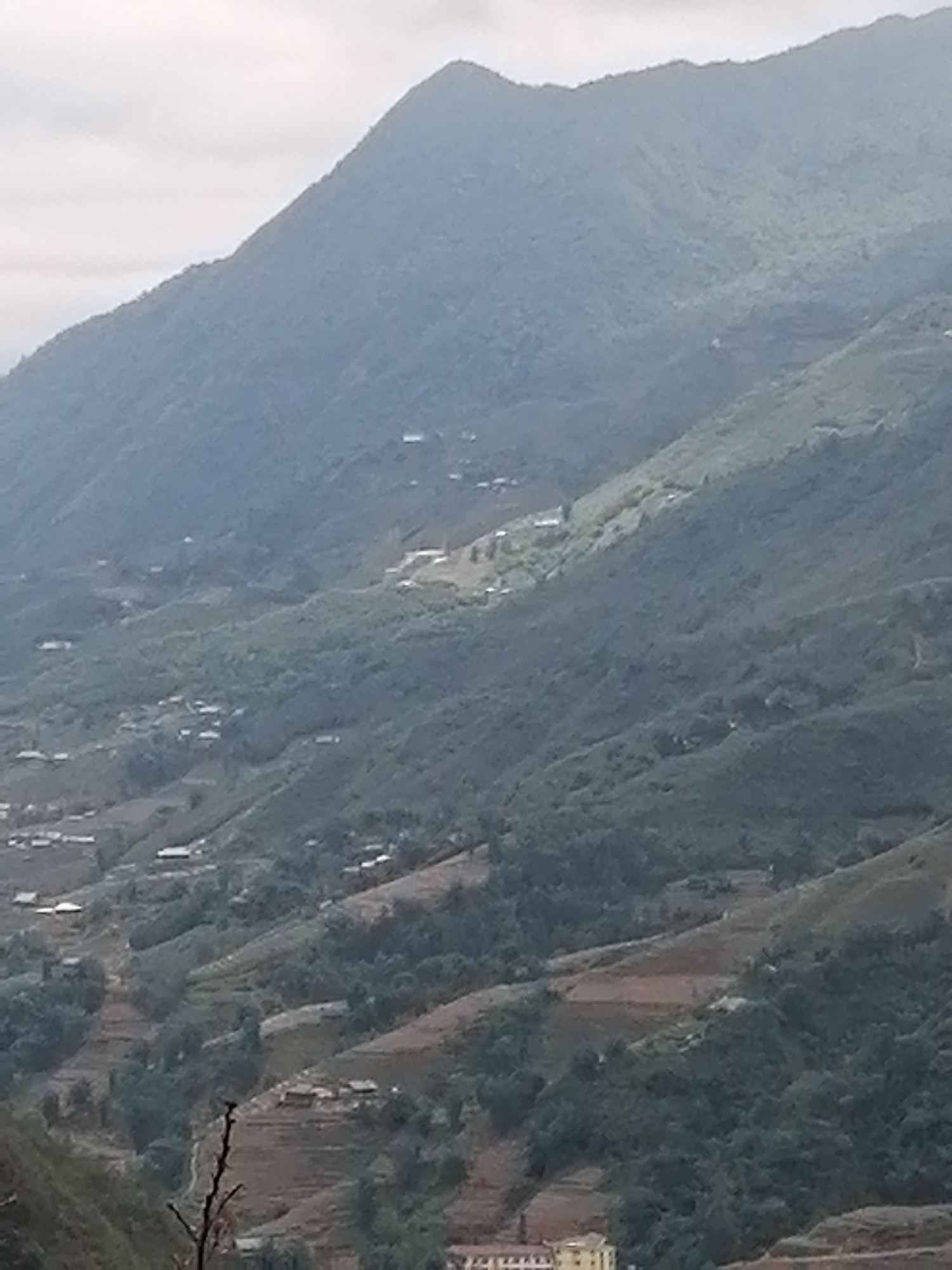 The image shows a hilly landscape, likely in Sa Pa Ward, Vietnam. A large mountain dominates the background, fading into a slightly overcast sky. The slopes of the mountain are terraced with vegetation and what appear to be cultivated fields. Small buildings and settlements are scattered along the hillsides, indicating human habitation. The foreground features a lower section of the terrain with denser foliage and the suggestion of buildings or structures at the very bottom.