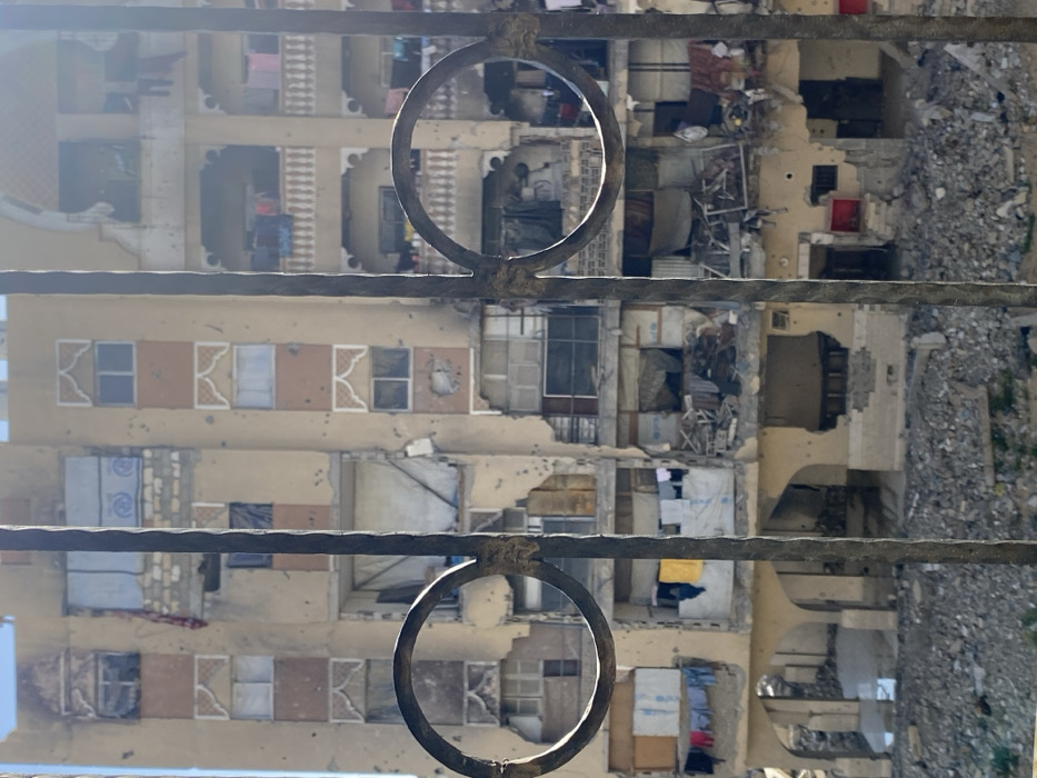 The image depicts a multi-story residential building in Khan Yunis, Palestinian Territory, viewed through rusted metal bars featuring two prominent circular elements. The building, constructed with tan and orange-brown facade panels and decorative arched window frames, exhibits severe structural damage. Numerous pockmarks and holes are visible across its exterior, indicative of bullet or shrapnel impacts. Windows are largely shattered or absent, with some openings covered by tarps or fabric. Balconies with ornate white balustrades are present on the right side, some with laundry hanging. Exposed interiors reveal significant debris and collapsed elements, particularly on the lower floors. A blue and white UNRWA tarp is visible on a lower-mid floor. The ground level in front of the building is covered by a dense field of rubble and concrete debris. Bright daylight illuminates the scene. No individuals are visible.