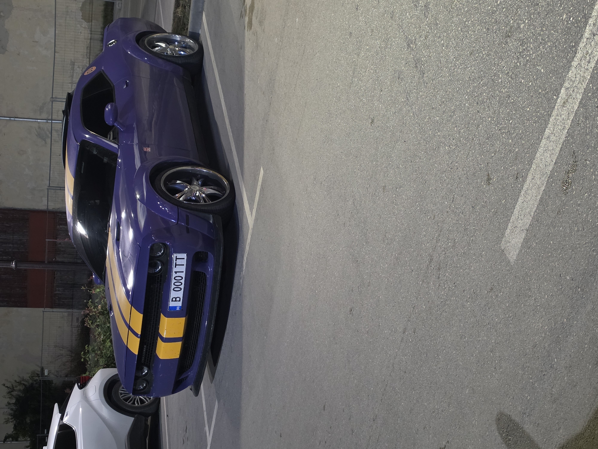 A purple Dodge Challenger with yellow racing stripes is parked in an outdoor parking lot. The car's license plate reads "B 0001 TT". A white SUV is parked to the left of the Challenger. The parking lot is paved with asphalt and marked with white lines. In the background, there is a fence and a building with a red roof. The scene is dimly lit, suggesting it is nighttime or dusk. The location is confirmed to be Varna, Bulgaria based on the license plate format.