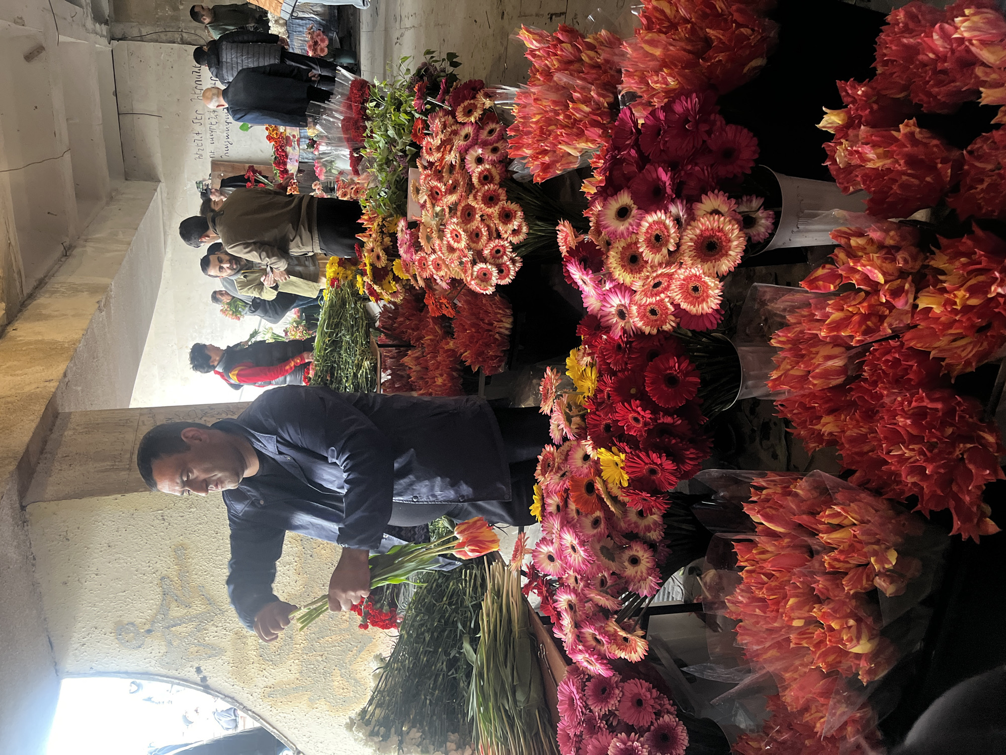 The image captures a bustling flower market in Yerevan, Armenia. A middle-aged man in a dark blue jacket carefully selects flowers, surrounded by vibrant displays of red, pink, and orange gerbera daisies and tulips. Other people are browsing and making purchases in the background. The market is situated indoors, likely within a building or tunnel structure with concrete walls. There appears to be some text or graffiti on the wall behind the man.