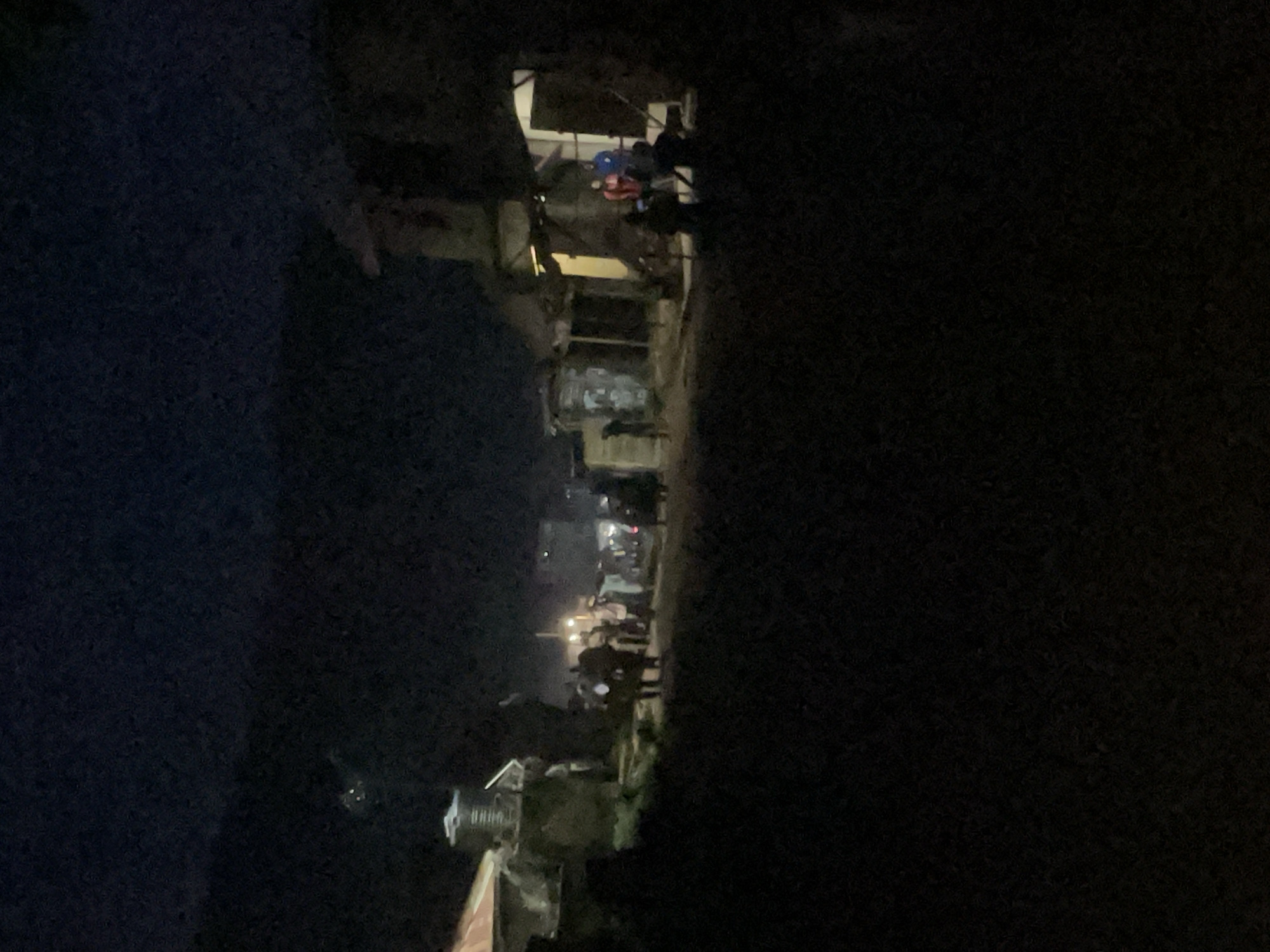 The image depicts a dimly lit outdoor scene at night, likely in a residential or commercial area within the city of Lokoja, Nigeria. Several individuals are visible, clustered in small groups along a street or pathway. Buildings with rudimentary structures and open doorways are present on both sides of the path. The lighting suggests the presence of artificial illumination from within some of the buildings or from streetlights, casting faint glows. The overall atmosphere appears to be that of a gathering or daily activity taking place after dark in a developing urban setting. The foreground is dominated by dark, indistinct shapes, possibly vegetation or uneven terrain.