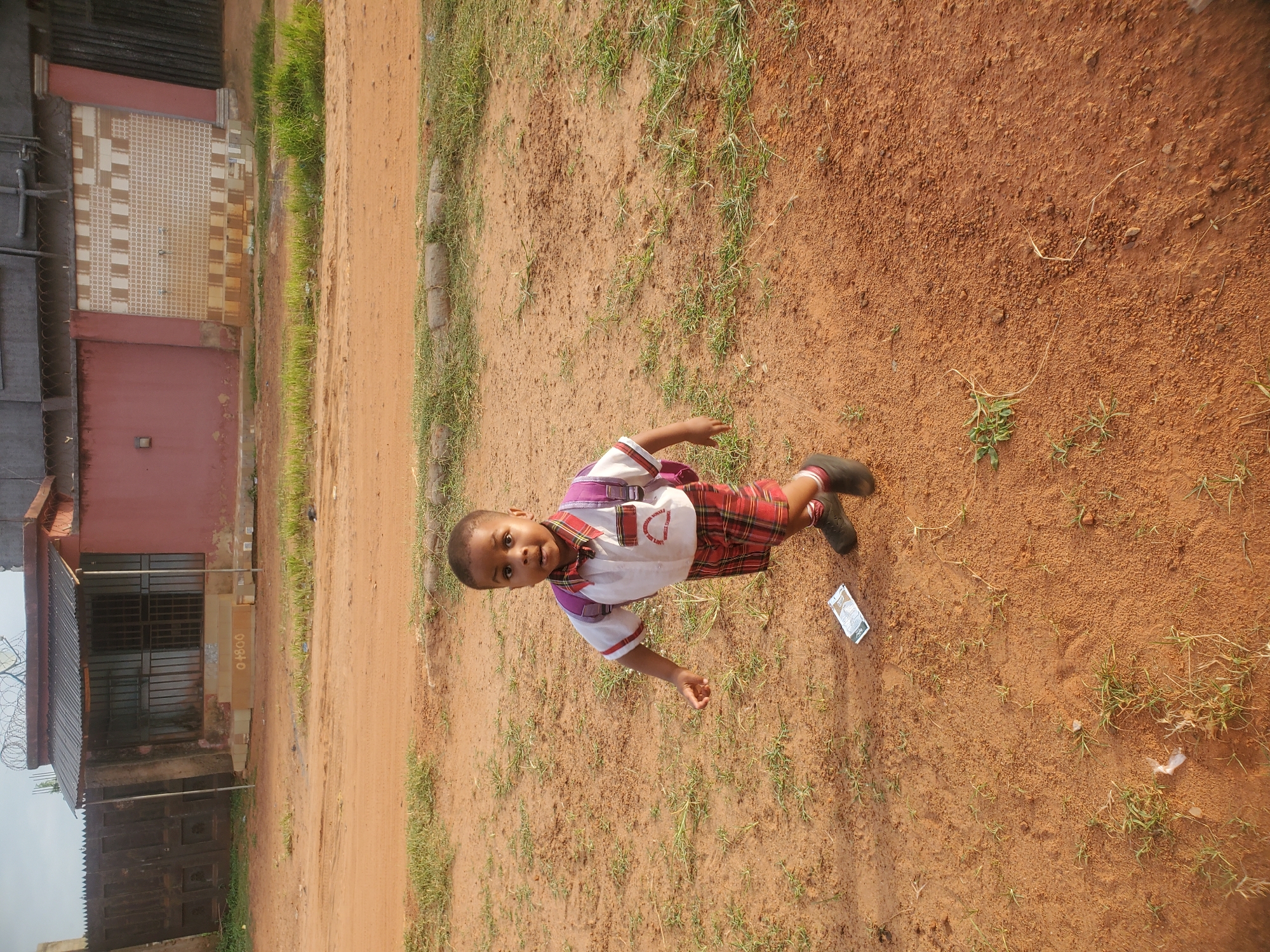 A child stands on a dirt patch next to a road. The child is wearing a school uniform: a white collared shirt, plaid red and brown shorts, and black shoes. A small backpack is slung over the child's shoulder. A building with a red facade is visible to the left of the frame. The ground is covered in red dirt with sparse patches of grass. A wrapper lies on the dirt next to the child. The location is Ukwu-Abwa, Nigeria.
