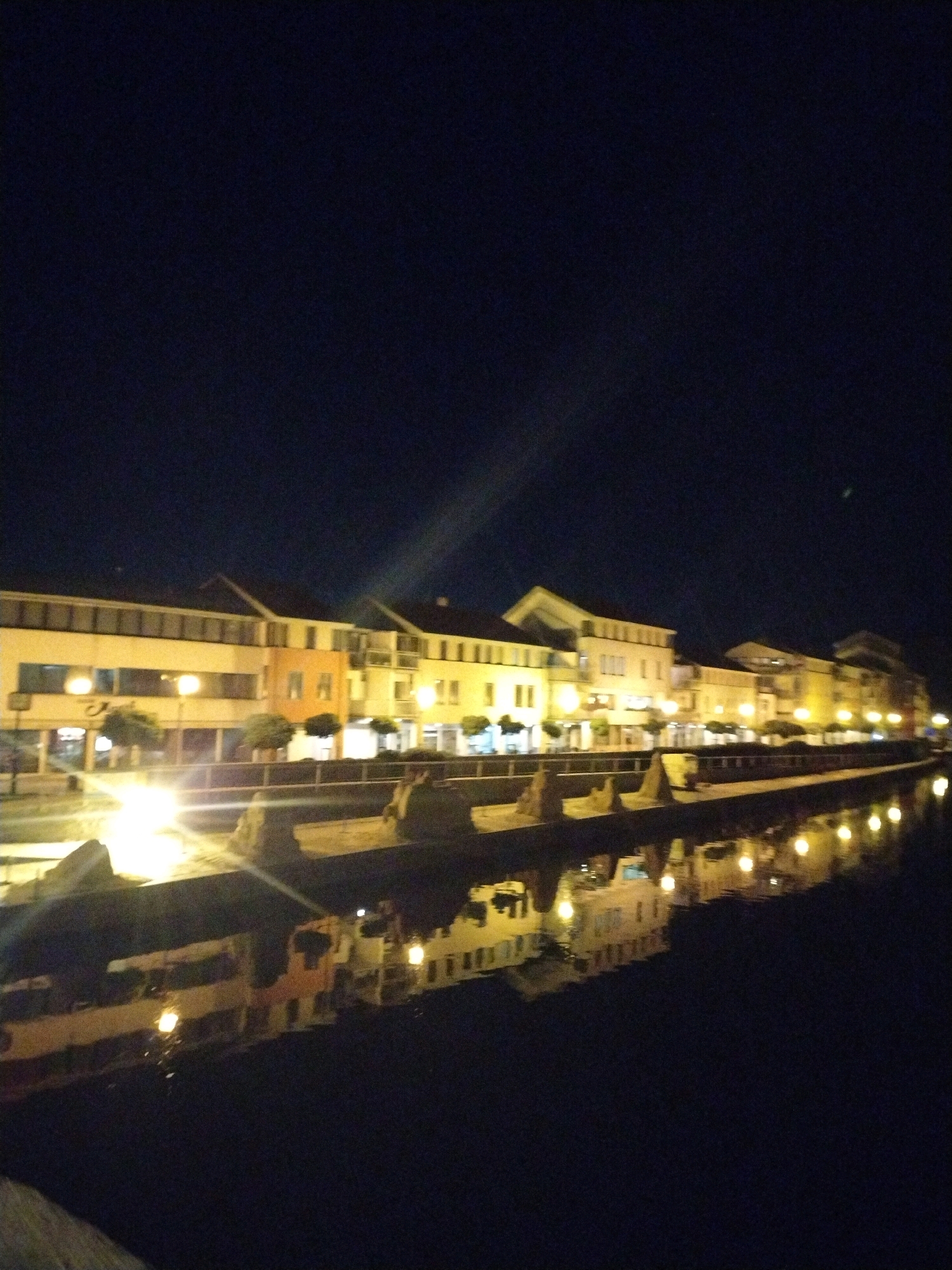This image depicts a nighttime scene along a river in Písek, Czechia. On the far bank, a row of buildings with illuminated windows lines the waterfront. The architecture appears to be modern, with multiple stories and large windows. The lights from the buildings reflect onto the dark water of the river, creating shimmering patterns. In the foreground, there is a dark embankment, possibly a riverwalk or path, with some indistinct structures or seating. The sky above is a deep, dark blue, characteristic of nighttime. There are no visible people actively engaged in actions within the frame, though the presence of seating suggests potential for public use. The overall impression is one of a tranquil, illuminated urban waterfront at night.