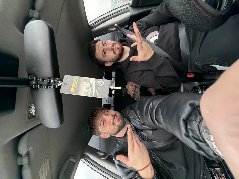 The image depicts the interior of a vehicle, seemingly parked or stopped, with three male occupants and an apparent fourth person taking a selfie. The location is Nevsha, Bulgaria, with a large body of water visible through the windows, suggesting a coastal or lakeside setting.
In the front passenger seat, a male with dark hair and a beard, wearing a black jacket, is smiling at the camera and making a horizontal pointing gesture with his right hand. A red beaded bracelet is visible on his left wrist. The driver, also a male with dark hair and a beard, is seated to the right, wearing a black "The North Face" hoodie and a seatbelt. He has a quizzical expression and holds his left hand open with the palm facing up. In the rear middle seat, a third male is seen sleeping or resting, with his head against the seatback and partially covered by a textured beige or light brown fabric.
The vehicle's interior features a gray headliner and sun visors. An "AREON QUALITY PERFUME" air freshener, depicting a classic car and cityscape, hangs from the rearview mirror by a black beaded chain. A black beverage can is visible in the center console between the front seats. A bare forearm with a metallic watch occupies the immediate foreground, indicating the selfie perspective.