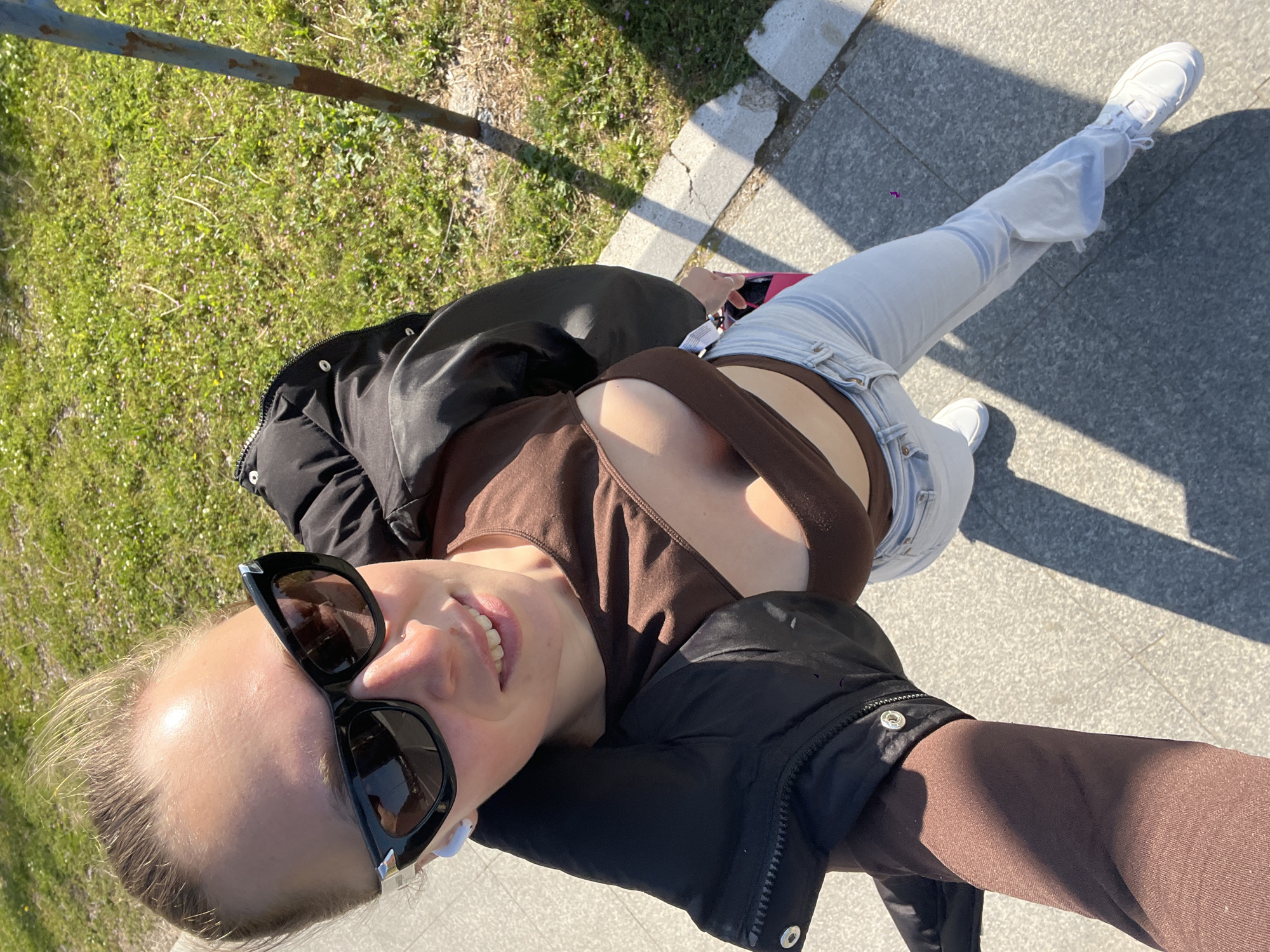 A young woman is standing on a gray paved sidewalk beside a patch of green grass, possibly in the city of Boreti, Montenegro. She is wearing a brown, cropped top, light blue jeans, and white sneakers. Over the top, she has a black puffer jacket. She is also wearing black sunglasses and has blonde hair pulled back. She is likely taking a selfie, as her arm is extended, and her face is centered in the frame.