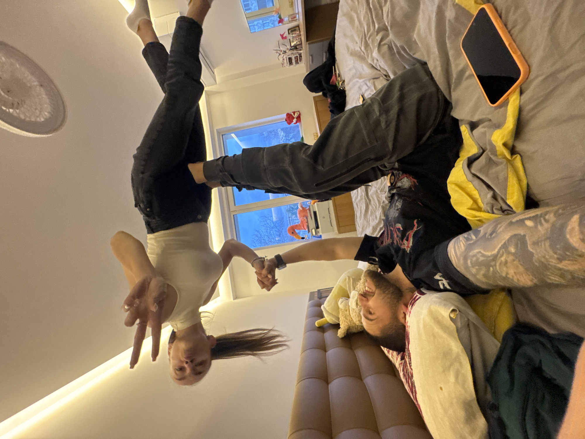 Two people are positioned in what appears to be a bedroom. A woman with dark hair is upside down, holding onto a man's arm. She is wearing a white top and dark pants, and her legs are extended upwards. She is making a peace sign with her left hand. The man is lying on a bed, wearing a black t-shirt and grey pants. The bed has grey linens and an orange-cased phone is visible. There is also a stuffed animal on the bed. In the background, a window reveals an outdoor scene, with what appear to be winter trees. There is an object outside resembling a flamingo on the window. The room is lit with artificial light. The location in the city of Sofia, Bulgaria cannot be verified with the given information.