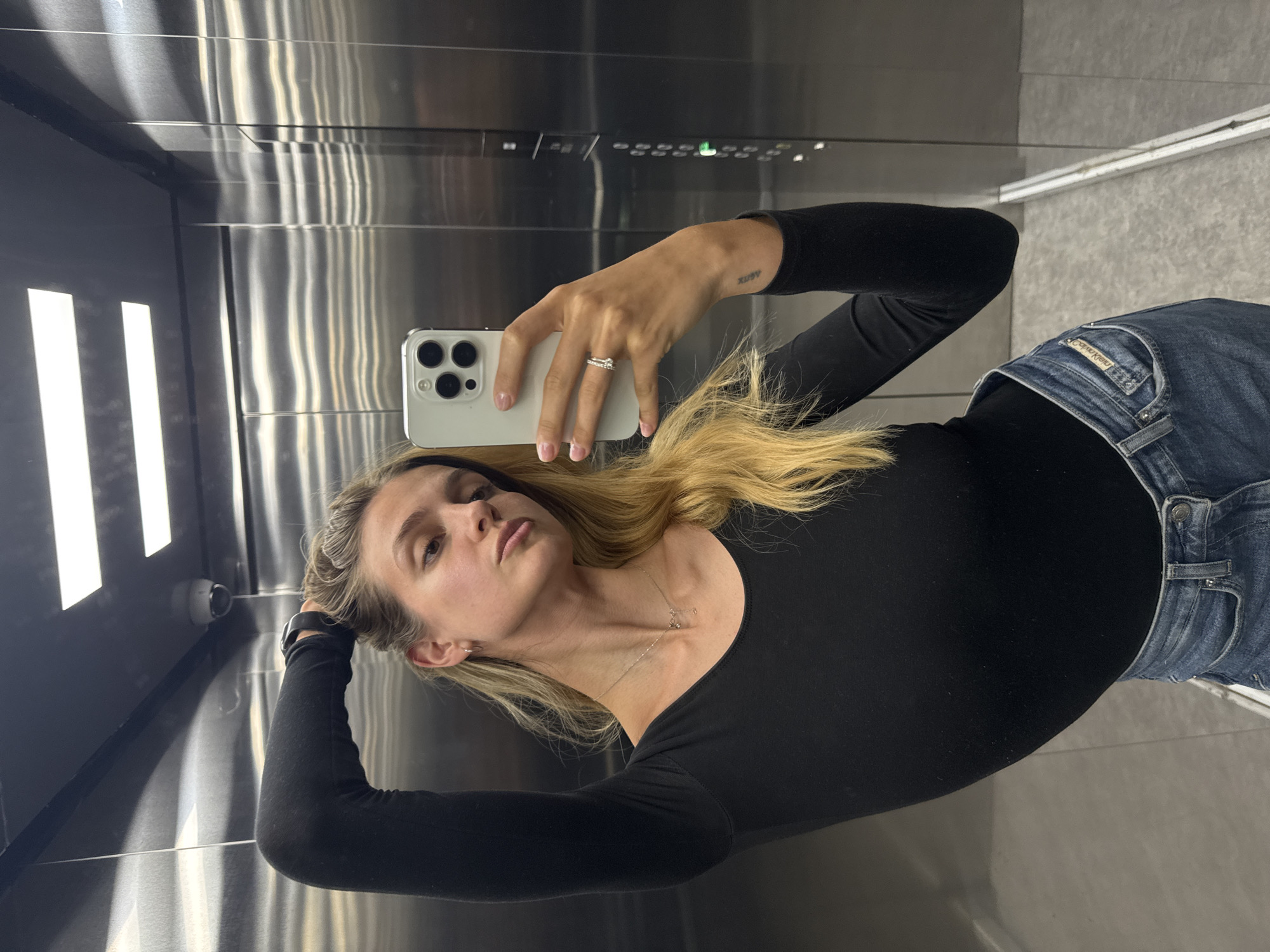 A woman is taking a selfie in an elevator. She is holding a white smartphone in her right hand, showing off a ring on her finger. She is wearing a black long-sleeved top and blue jeans. She has blonde hair and is wearing a necklace. The elevator has a metallic interior. A security camera is visible in the upper left corner. The elevator control panel is above her right hand. The floor is a light gray color. The location is Unknown, Unknown.