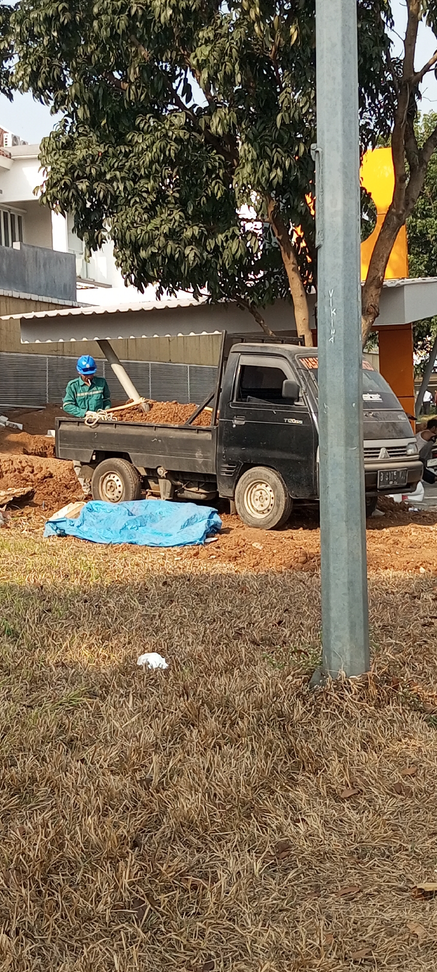 In Lambangjaya, Indonesia, a construction scene unfolds. A black pickup truck is parked on a dirt lot, its bed filled with soil. A construction worker, wearing a blue hard hat and a green uniform, stands in the truck bed, holding a cigarette. Nearby, a blue tarp is spread on the ground, partially covering what appears to be an object. Dry grass covers the foreground, with a small white object visible in the grass. A utility pole stands in the foreground to the right of the truck. In the background, a building and trees can be seen.