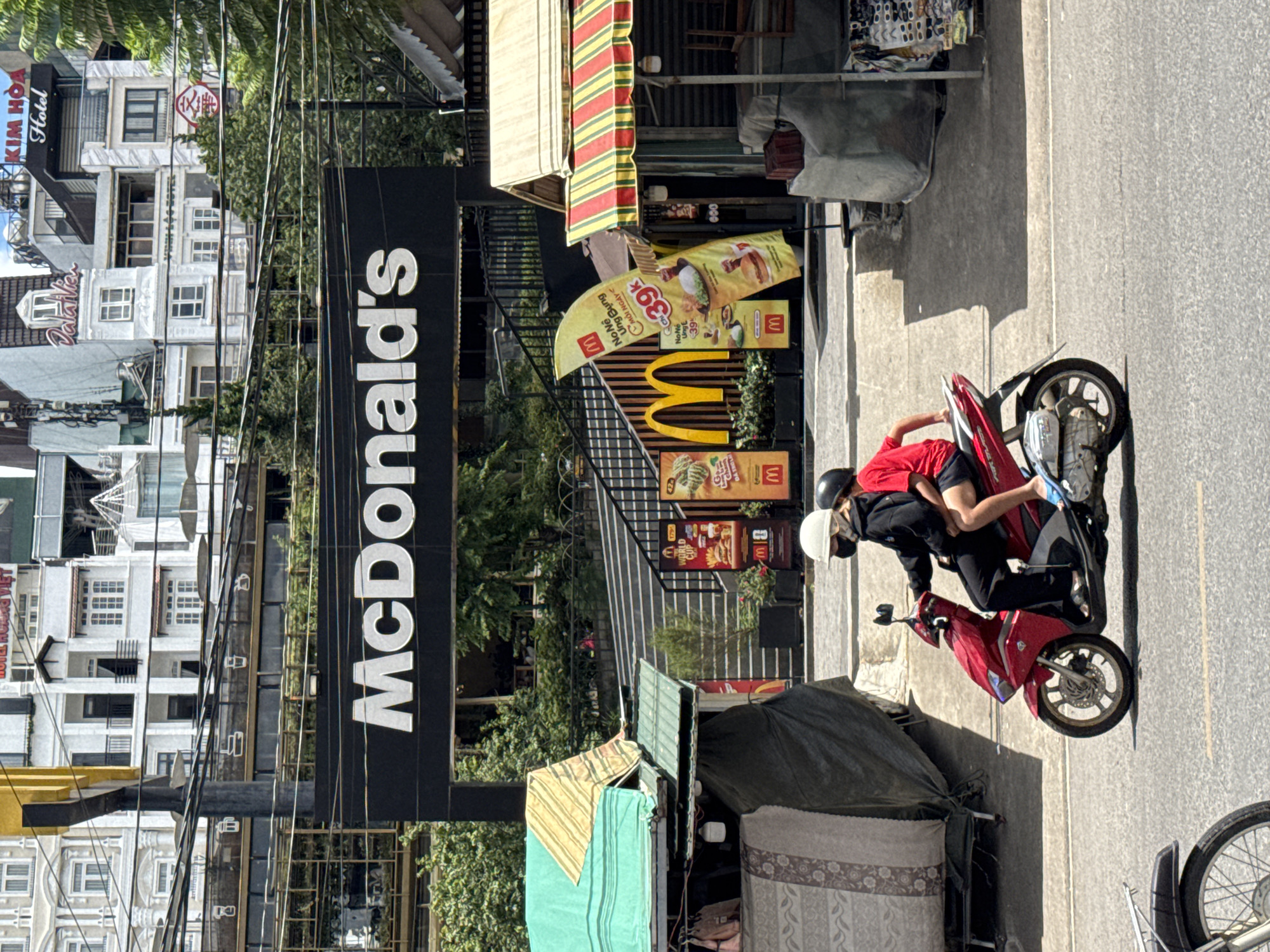 A red scooter with two occupants rides down a street in Da Lat, Vietnam. The driver wears a black jacket, white helmet, and face mask. The passenger, wearing a red t-shirt and black shorts, sits behind the driver. In the background, a large McDonald's sign is visible above a set of stairs. Several market stalls and buildings line the street.