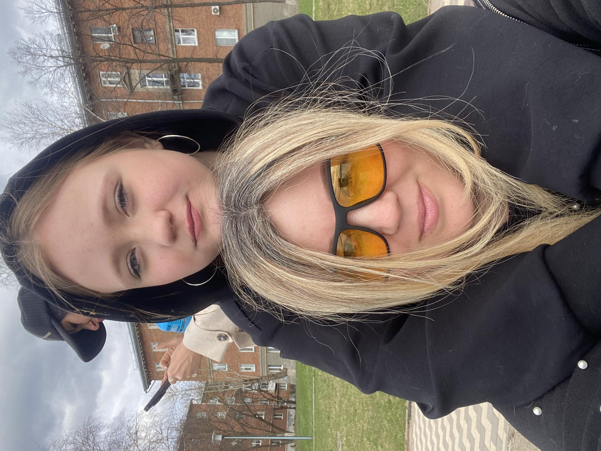 Two women are photographed outdoors in what appears to be a city setting. The younger woman, on the left, wears a black hooded sweatshirt, has light skin, and large silver hoop earrings. The other woman on the right wears orange-tinted sunglasses and has light blonde hair with gray roots. Both appear close to the camera, perhaps in a selfie. The woman on the right has her arm around the shoulders of the woman on the left.
In the background, a brick building with multiple windows and an air conditioning unit can be seen. There is also a grassy area, bare trees, and a paved walkway with a geometric pattern. The location appears to be in the city of Votkinsk, Russia, based on visual landmarks. Another person can be seen holding a water bottle and a black stick or pole with a tan or off-white jacket.