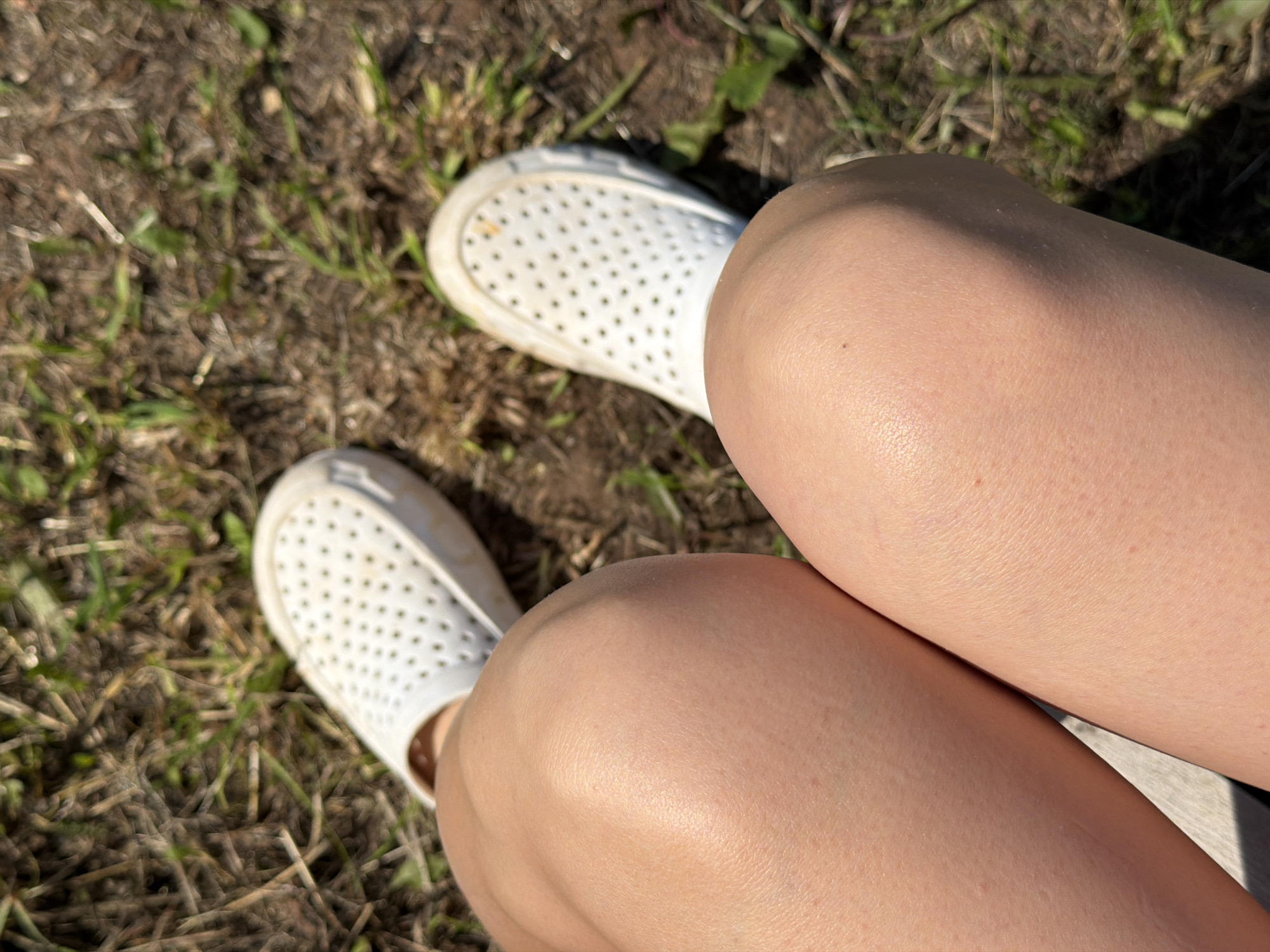 A close-up, top-down view shows a person's bare legs and feet, clad in white perforated slip-on shoes, resting on dry grass and dirt. The knees are prominent in the foreground, with one leg bent at the knee and the other extended slightly. The shoes appear to be made of a rubber-like material, with numerous small circular holes across their surface. The ground is covered with a mixture of dry grass, straw, and some scattered green shoots, suggesting an outdoor, possibly unkempt, setting. The lighting indicates a bright, sunny day. The image does not contain information about the city of Chaikovsky, Russia.