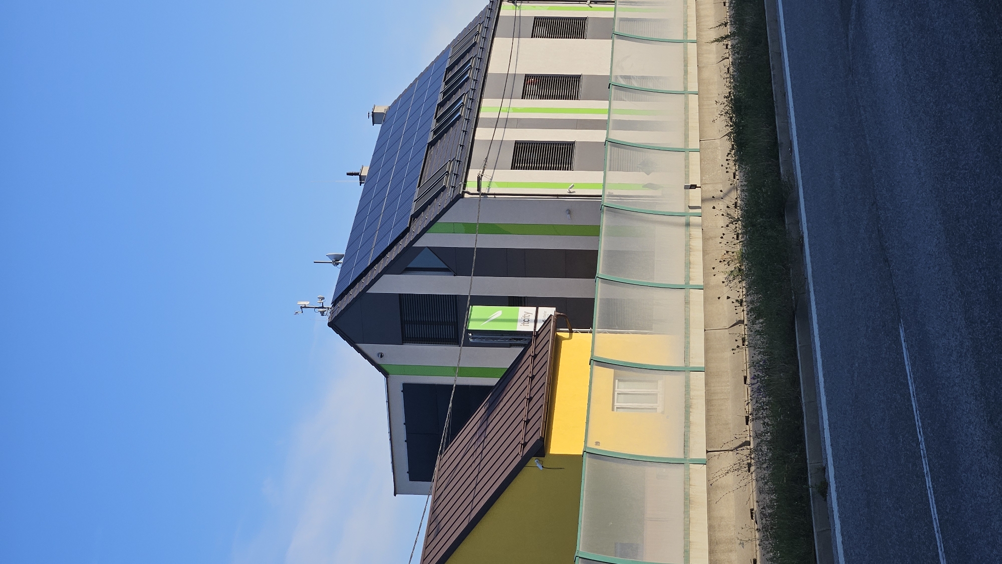 The image shows a modern building with solar panels on its roof, located in Prešov, Slovakia. The building features a striped facade with green and grey vertical lines, and a prominent green banner with the text "itcity" and "energia". To the left of this building is a smaller, yellow building with a brown roof. A transparent noise barrier, supported by green metal posts, separates the buildings from a road in the foreground. The sky is clear and blue. The scene appears to be an exterior view of a commercial or industrial area.