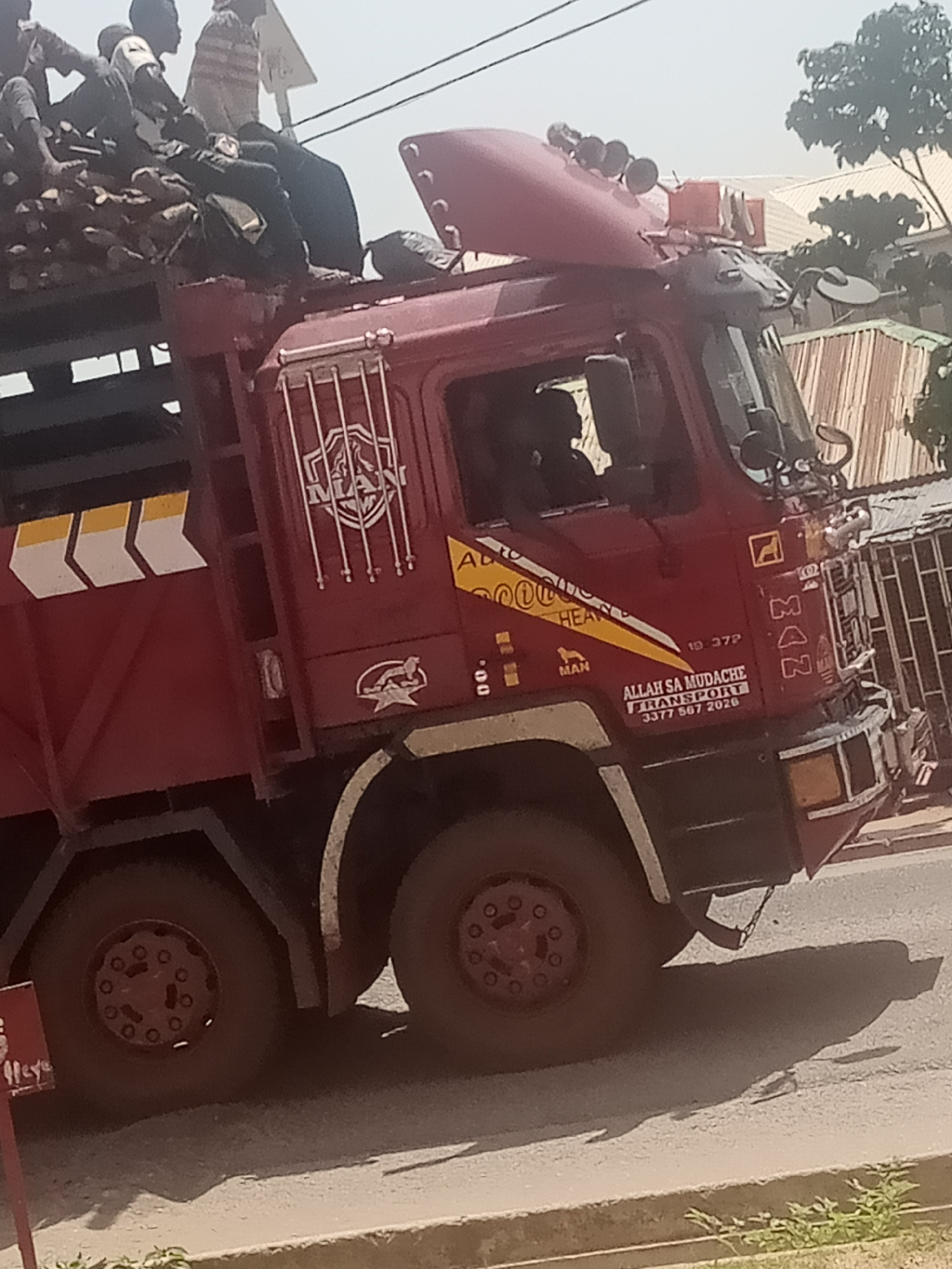 The image shows a maroon MAN truck overloaded with firewood and people. Several individuals are seated on top of the firewood, with some appearing to be standing. The truck is in motion, suggesting it is either on its way to a destination or has just departed.
The scene appears to be outdoors, possibly on a road in Jalingo, Nigeria. The lighting suggests it is daytime, with a bright sky and shadows cast on the ground. The presence of trees and buildings in the background gives a glimpse of the urban or semi-urban environment.
Notable details include the brand "MAN" prominently displayed on the truck, along with the text "ALLAH SA MUDACHE TRANSPORT" and a phone number "3377 507 2020". There are also yellow and white chevron markings on the side of the truck's cargo area. The overall impression is one of a busy and practical scene, likely related to transportation of goods and people in a region where such methods are common. The people on the truck appear to be engaged in their journey, with no overtly discernible emotions.
