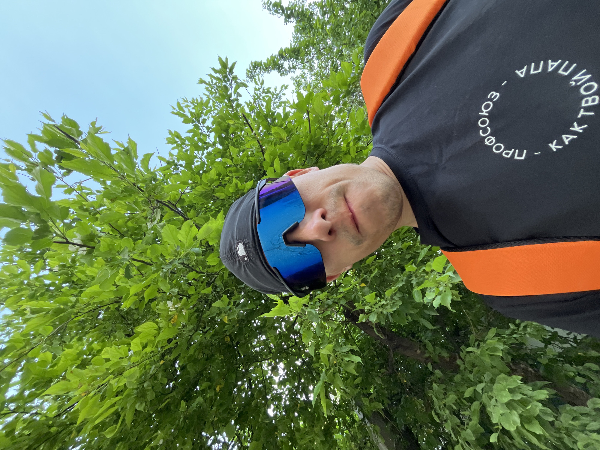 A low-angle, outdoor selfie shows a man wearing a black cap and mirrored blue sports sunglasses. The man has a fair complexion and is wearing a black shirt with orange straps. White text in a circular pattern is visible on the shirt. Green foliage fills the background, including tree branches and leaves, suggesting a park or forested area. The sky is visible in the upper left corner. The photograph was taken in the city of Chaikovsky, Russia.