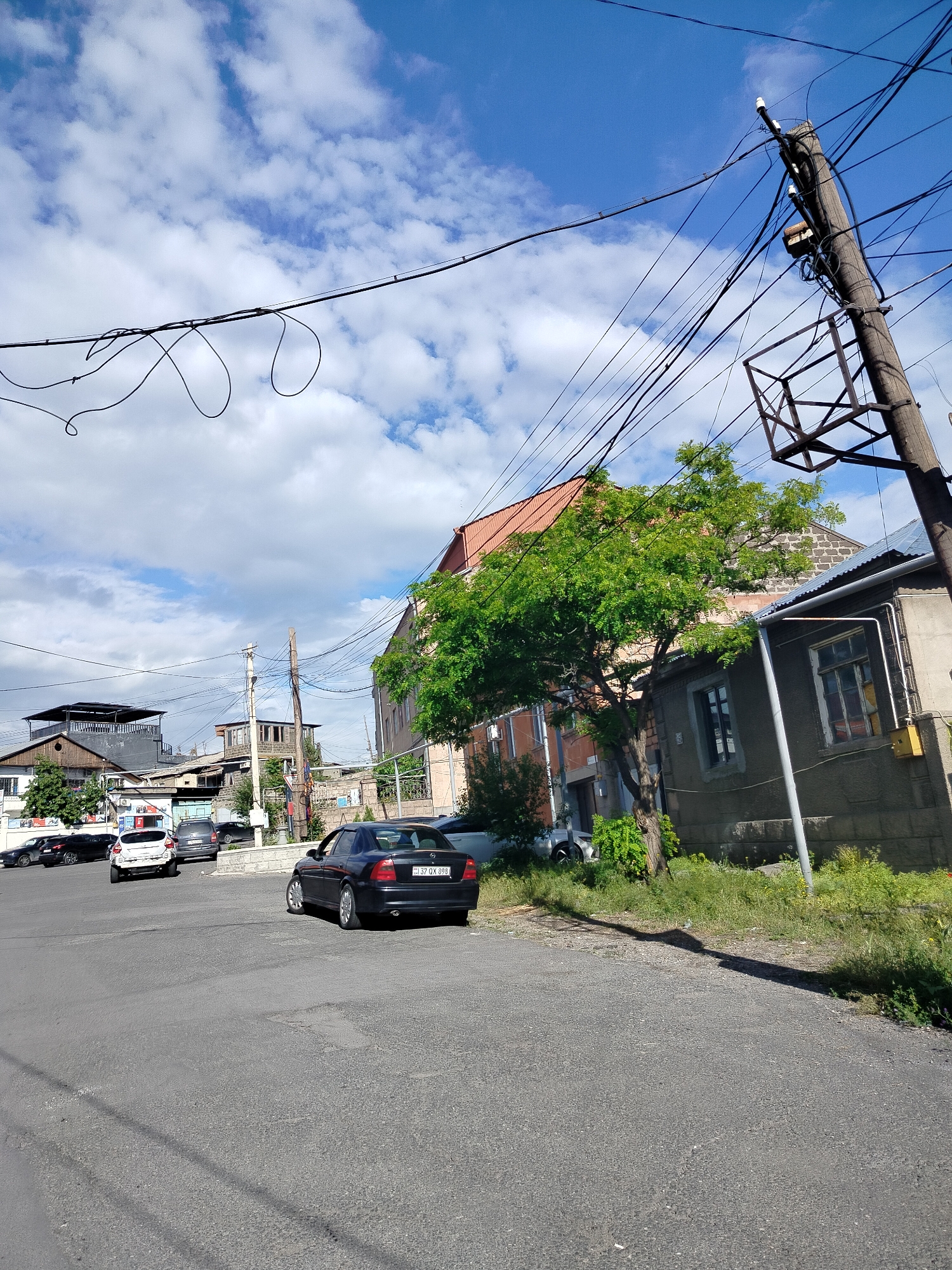 The image depicts a street scene in Yerevan, Armenia, on a partly cloudy day. A black Opel car, license plate "32 QX 899", is parked on the right side of the road, facing away from the camera. Several other cars, including a white one, are parked further down the street in the background. A wooden utility pole with attached electrical equipment and wires dominates the right side of the frame. Buildings of varying heights and architectural styles line the street, including a single-story structure with a gray exterior and a building with a red tile roof. A medium-sized tree stands next to the gray building. The road surface is asphalt. The sky is blue with scattered white clouds. The overall lighting is bright and sunny.