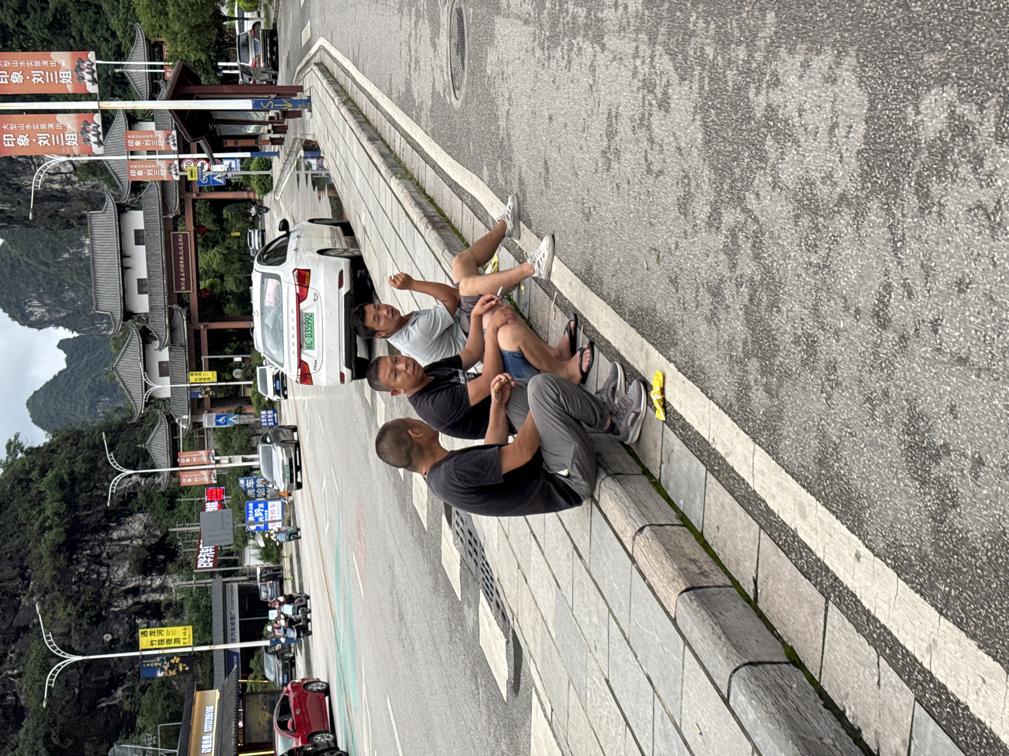 Three men are sitting on a low wall bordering a road in Guilin, China. The man closest to the viewer is wearing a black t-shirt and gray pants. The second man wears a black t-shirt and jean shorts, and the third man wears a white t-shirt and jean shorts. Cars pass by in the road beyond the wall. Buildings with traditional Chinese architecture are visible on the left side of the image, including billboards written in Chinese. Mountains can be seen in the background.