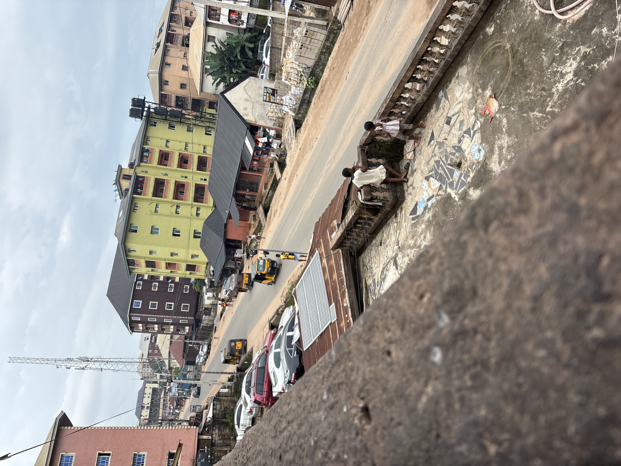 The image captures a street scene in Nkpor, Nigeria, under an overcast sky. In the foreground, two young children stand on a tiled balcony, one in a white dress, the other in a patterned top and shorts. A rusty, mottled concrete railing separates them from the street below. Across the street, a busy road is lined with buildings of various colors and heights, including a prominent yellow four-story building and a dark brown building. Several cars, including a red SUV, a white sedan, and a black sedan, are parked along the side of the road. Yellow auto-rickshaws, a common mode of transport in Nigeria, are also visible on the road, along with pedestrians walking and interacting. A large telecommunications tower stands tall in the background, with various antennas attached. Construction materials, including stacks of cement blocks and bags of sand, are piled on the side of the road near a small shop. A solar panel is installed on a corrugated metal roof in the middle ground. The overall impression is of a vibrant urban environment with elements of everyday life and development.