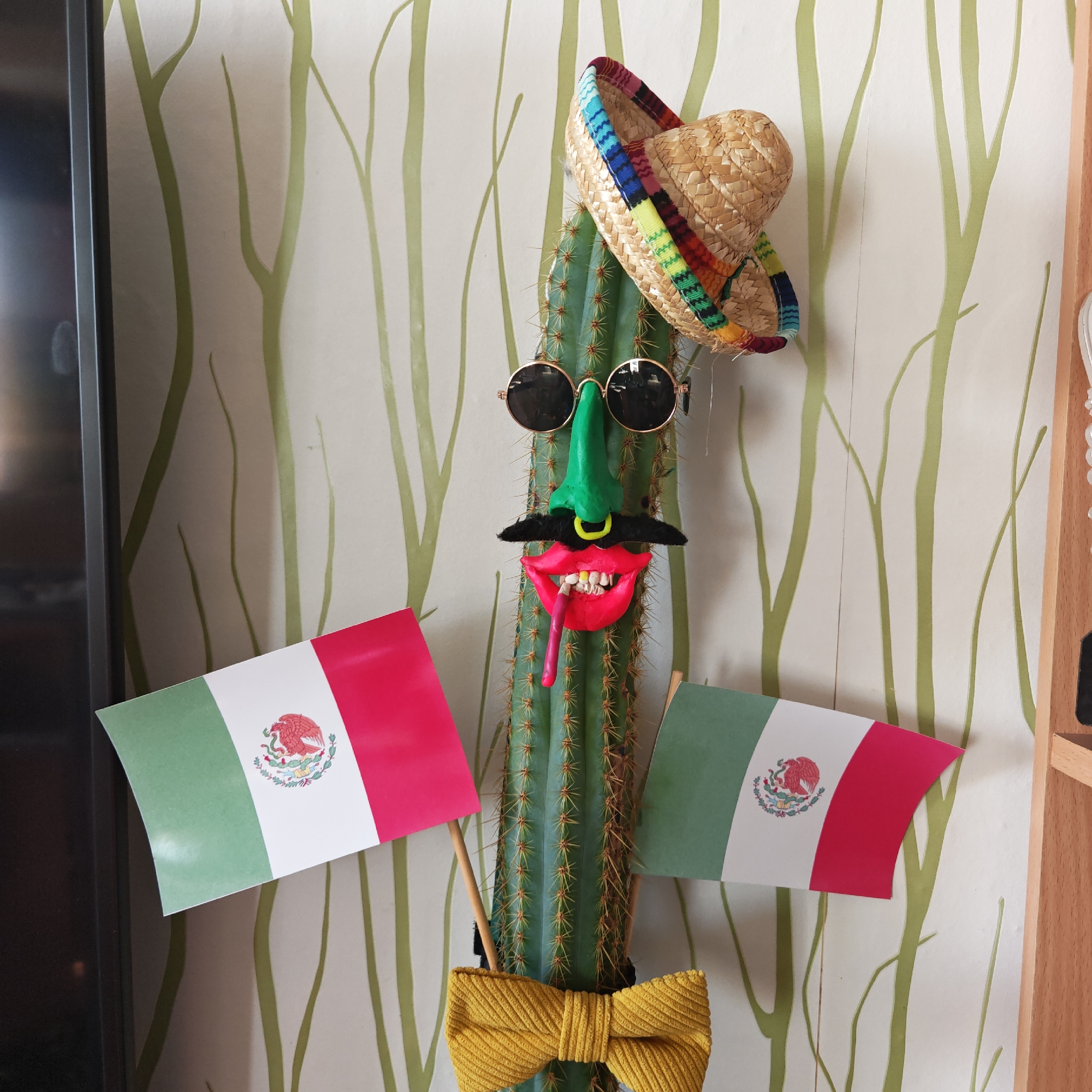 A cactus plant is decorated in a humorous, Mexican-themed manner. The cactus is adorned with a straw sombrero, round sunglasses, a large green nose, a black mustache, and a pair of bright red lips with exposed teeth and a red tongue. Two small Mexican flags flank the cactus on either side, and a yellow bow tie is tied around the base. The cactus stands against a wall covered in a white wallpaper with an abstract pattern of green vertical lines. Part of a television is visible on the left side, and part of a wooden shelf is visible on the right side. There is no indication that this picture was taken in Rostov, Russia.