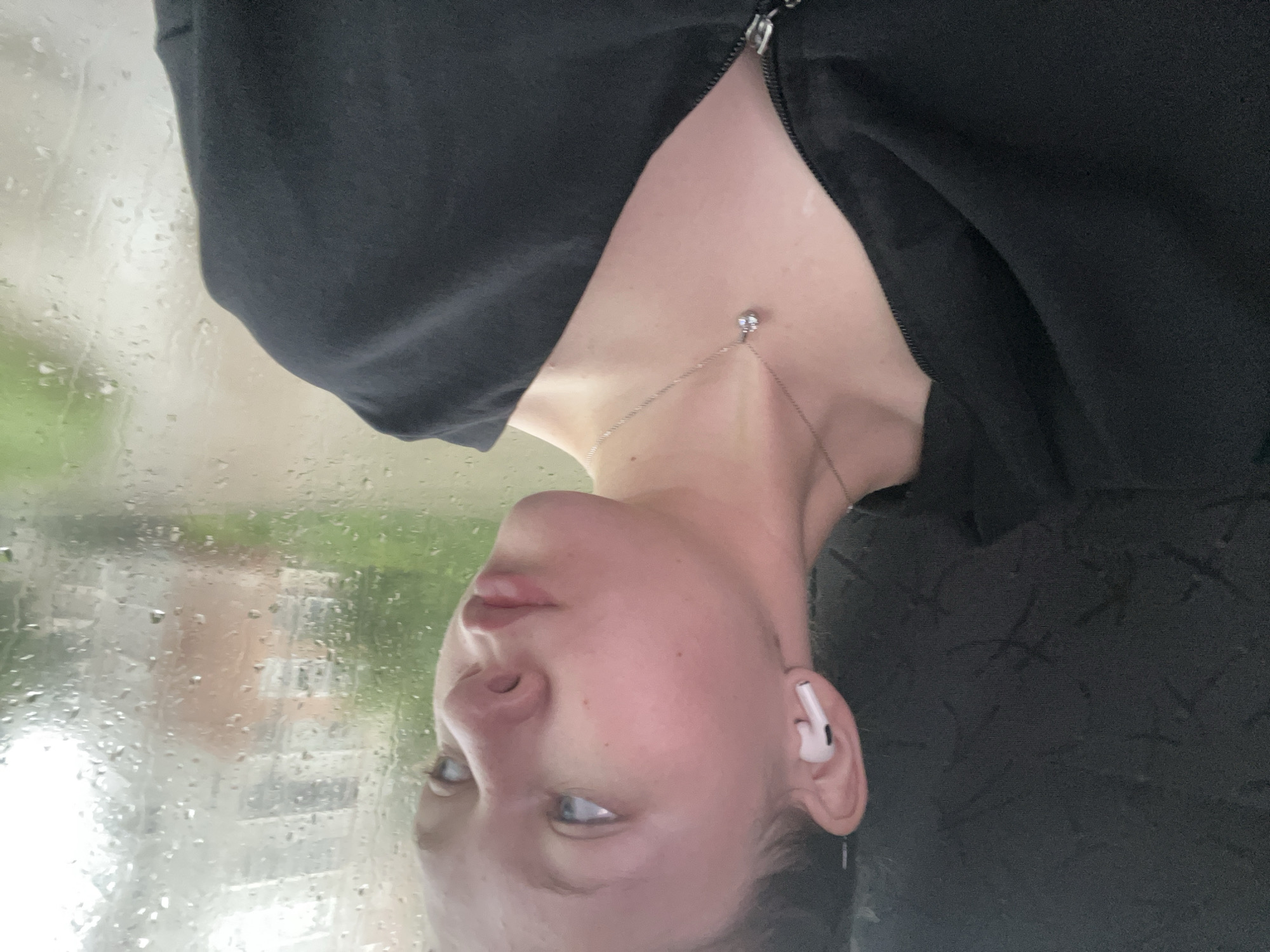 In Krasnokamsk, Russia, a young person with fair skin and light-colored eyes is captured in a close-up, profile shot. They are wearing white wireless earbuds in their left ear and a silver necklace with a small, clear pendant. The person is looking towards the right, with their mouth slightly open. They are wearing a black hooded garment, possibly a jacket or sweatshirt, with a visible zipper. The background is a window with raindrops on it, obscuring the view of buildings and greenery outside. The lighting suggests an overcast or rainy day.