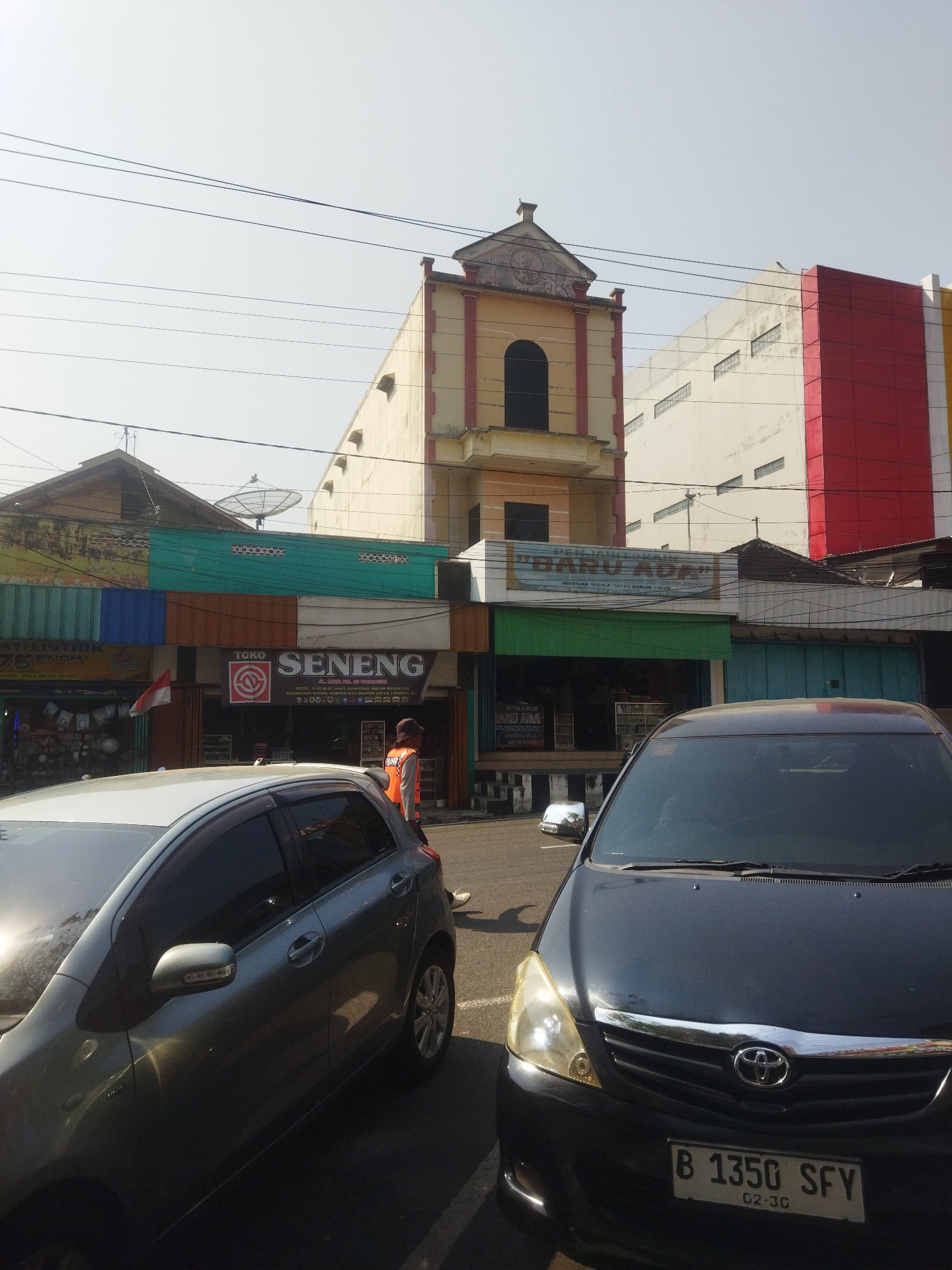 The image captures a street scene in the city of Wonogiri, Indonesia, bathed in bright daylight. In the foreground, a dark colored Toyota Innova is parked, its front end facing the viewer. To its left, a silver hatchback is parked, angled towards the left side of the frame. The backdrop features several commercial buildings. A prominent building with a cream-colored facade and red trim, topped with a decorative architectural element, stands tall. To its right, a large white building with red and yellow sections is visible. Below these structures, a row of shops is present, with awnings and signage. One sign prominently displays "TOKO SENENG". A person wearing an orange vest and a hat is seen standing in front of one of the shops, facing away from the camera. Several power lines crisscross the sky above the buildings. The street appears to be paved.