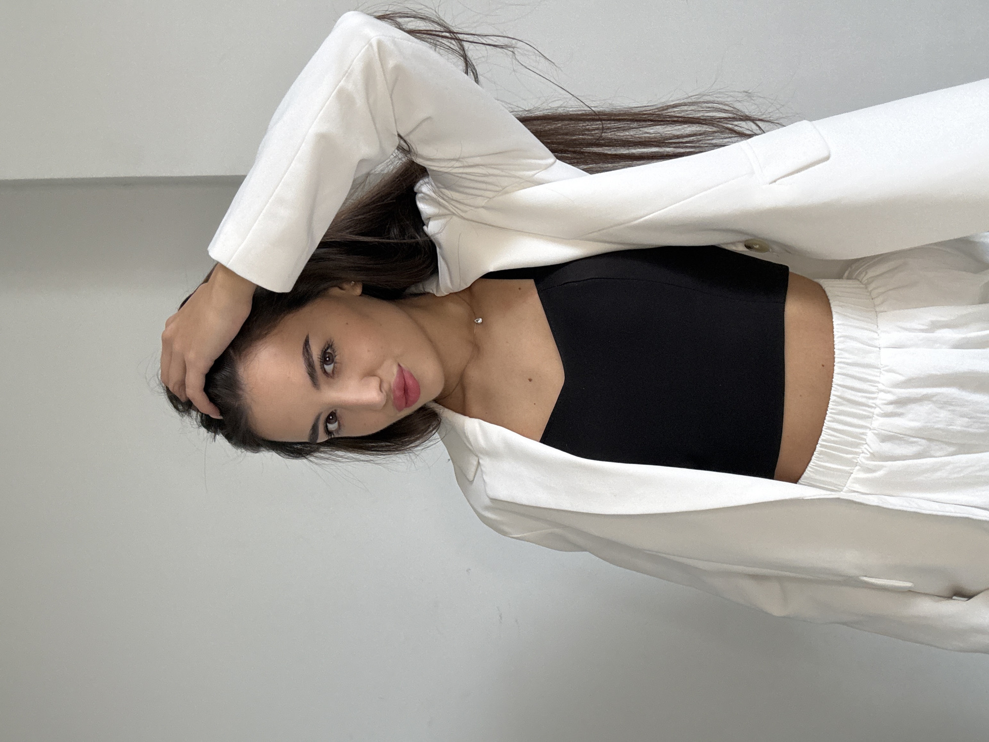 A young woman with long, dark hair is posing against a plain, light gray wall. She is wearing a white blazer over a black crop top and white pants. Her left hand is raised, touching the top of her head. She is looking towards the camera with her mouth slightly open, as if about to speak or smile. A delicate necklace with a small pendant is visible around her neck. The lighting is even, suggesting an indoor setting. There is no discernible indication of her location within Chișinău, Moldova.