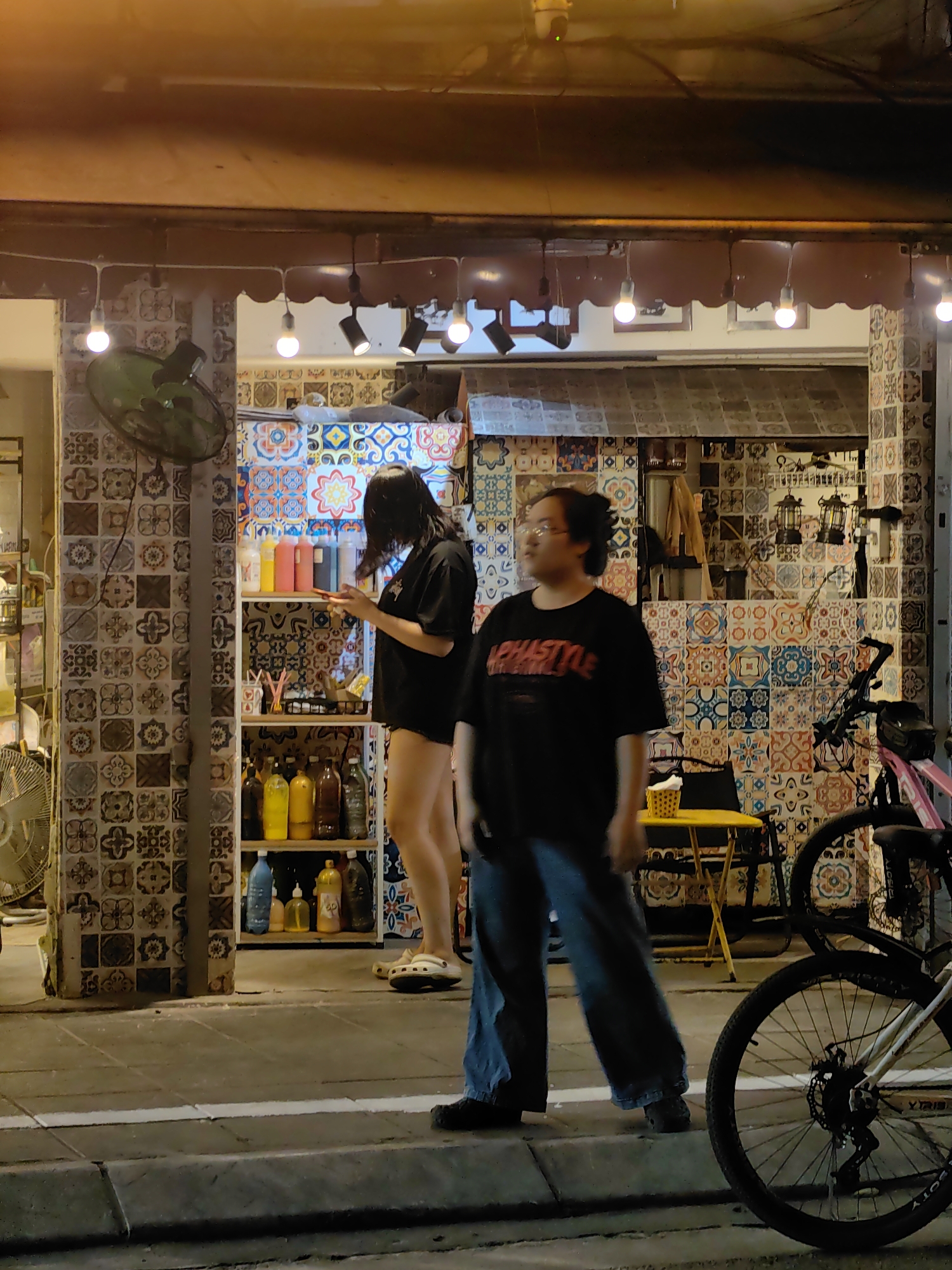 The image captures a scene in Hà Nội, Vietnam. A woman with dark hair, clad in a black t-shirt, is occupied with her phone in front of a shelf of colorful liquids. A man stands nearby, facing the camera, wearing a black t-shirt with lettering and loose blue jeans. The facade of the establishment, lined with patterned tiles, features a shelf with various bottles and containers. A yellow folding table and bicycle are visible on the right. The scene is illuminated by warm artificial light.