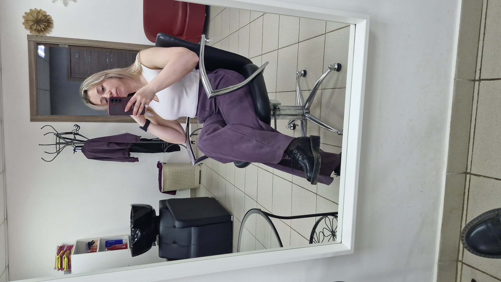 A reflection of a woman taking a selfie is visible in a mirror. The woman is seated in a salon chair. She's wearing a white tank top, purple pants, and black boots. She is holding a cell phone in her right hand. The reflection reveals a salon environment: a coat rack with hanging clothes, a hair washing station, and assorted salon supplies. The background is tiled. Based on the location provided, this scene is likely taking place in a beauty salon in Otchor, Russia.