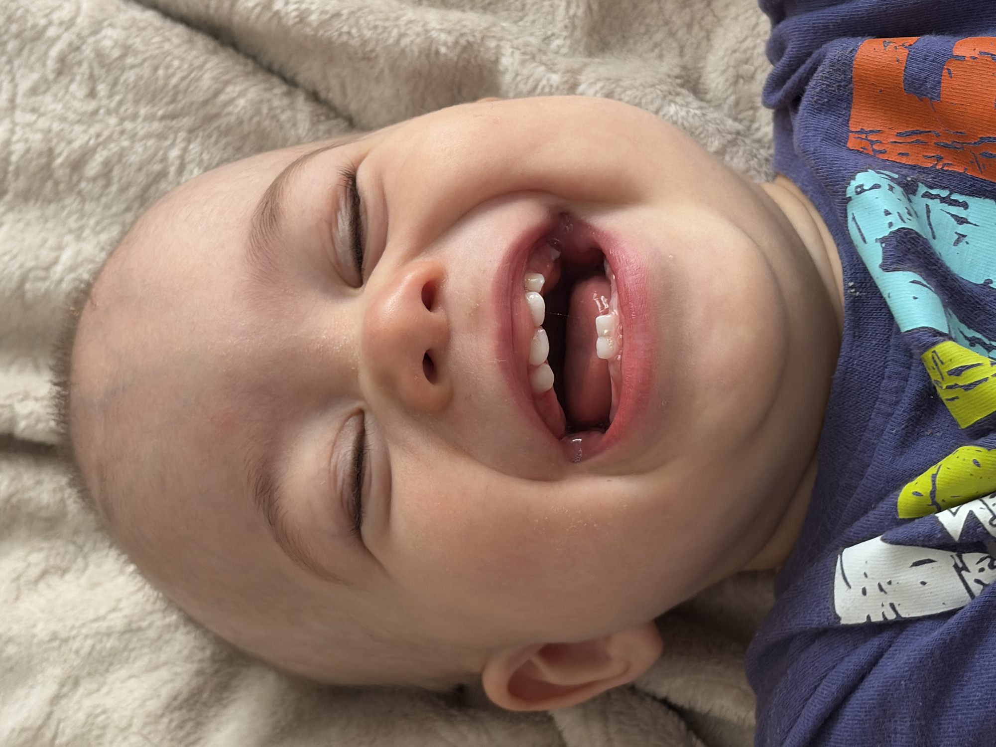 A close-up, angled overhead shot shows a baby lying on a beige textured surface, likely a blanket. The baby is fair-skinned with closed eyes. Its mouth is open, revealing several small, white teeth and a portion of its tongue. The baby wears a blue t-shirt with a colorful graphic design of orange, blue, and green. No other surroundings are visible to determine a location. There is no indication of the location being Chaikovsky, Russia.