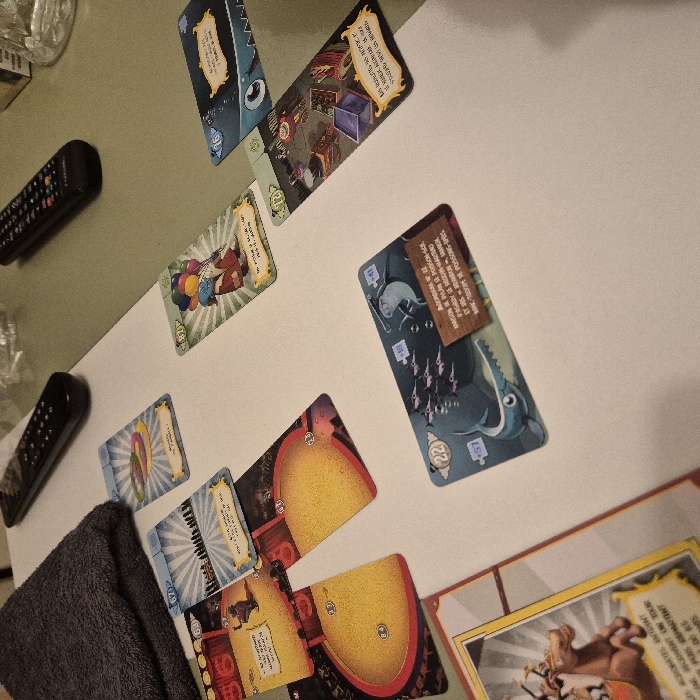 Several game cards are arranged on a white surface. A few cards depict illustrated scenes such as an acrobat, a blue shark swimming amongst a school of fish, and a room filled with objects. Text is visible on some cards, including phrases like "LES ACROBATES INTENT," "2 ON TOUR," and "+81 ASIA 21 KARUSION DE PYLAN POSSON-SCE." The cards are tilted and overlap slightly, creating a visually dynamic layout. Adjacent to the cards are a gray cloth and two remote controls.
